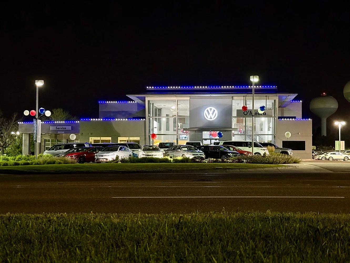 Night view of a Volkswagen car dealership with cars parked outside, blue and red decorations, and bright lights illuminating the building.