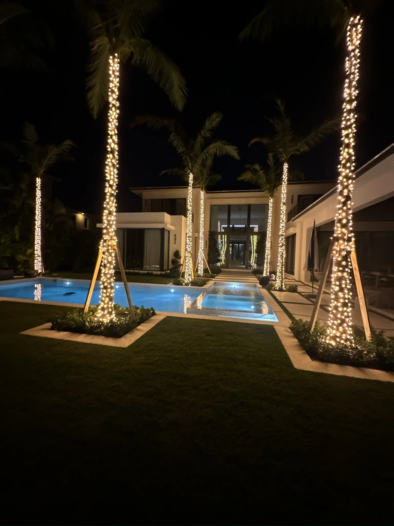Night view of a modern house with an illuminated swimming pool, poolside palm trees wrapped with string lights, and a pathway leading to the house entrance.