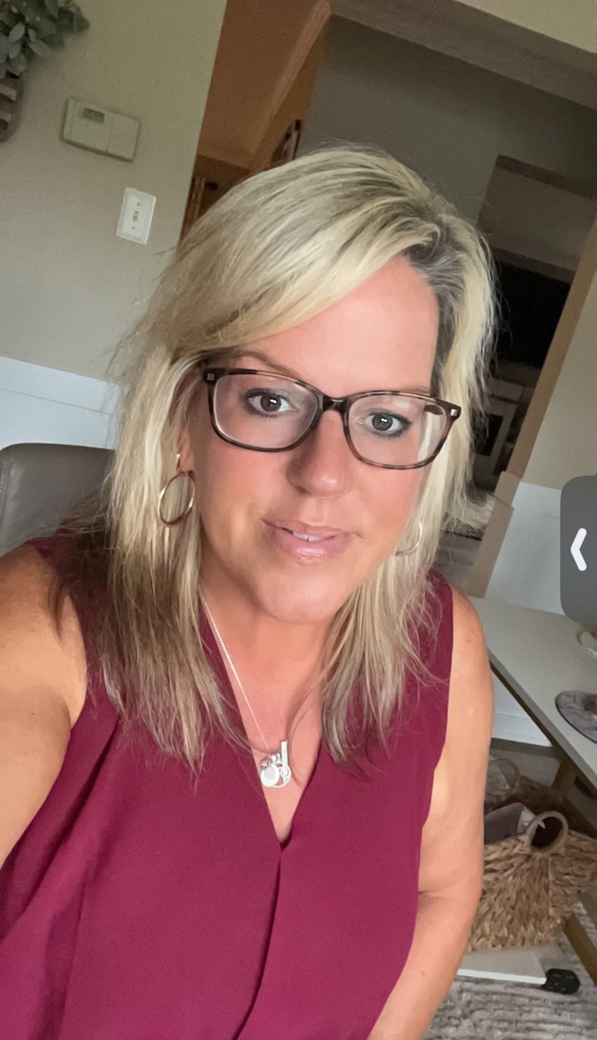 A woman with blonde hair, glasses, and hoop earrings wearing a sleeveless maroon top, sitting indoors with a kitchen visible in the background.