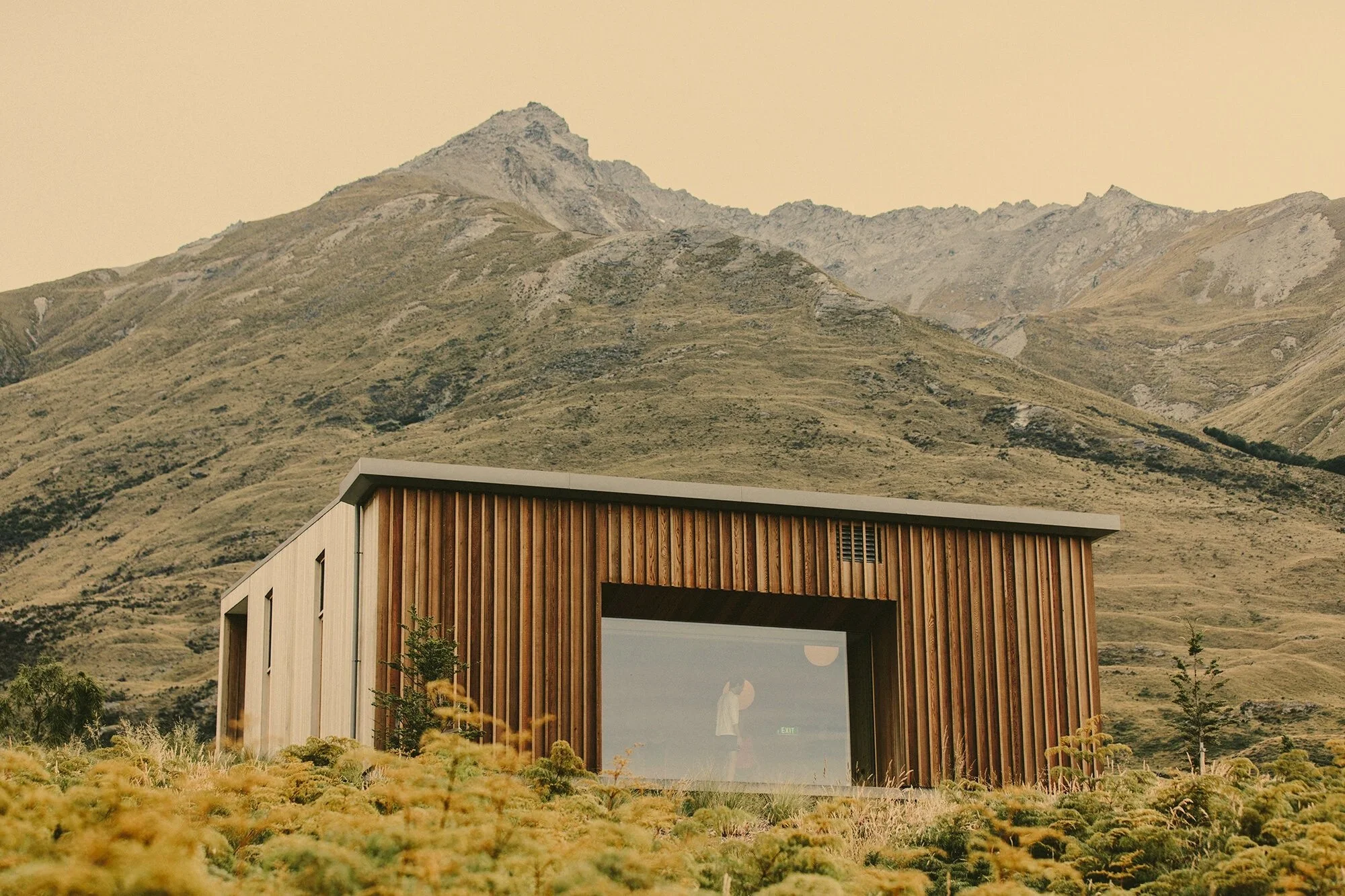 Modern building with wooden vertical slats and large glass window, set against mountainous landscape with dry grass and shrubs in the foreground.