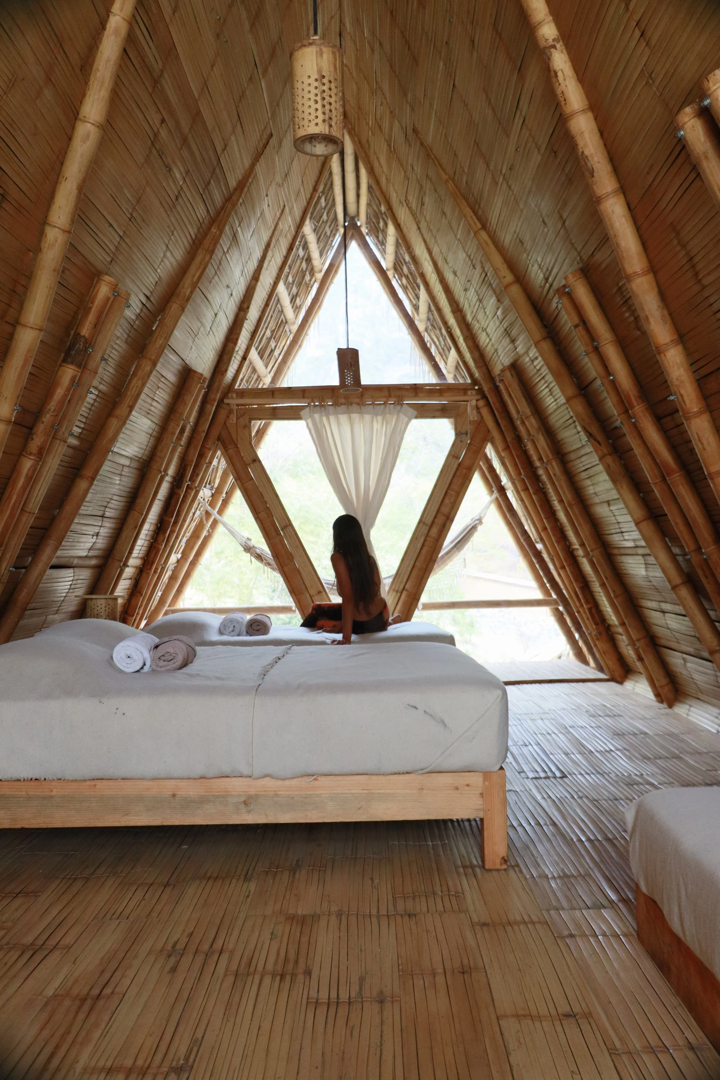 Interior de una cabaña de madera con una cama blanca, toallas enrolladas, cortinas blancas y ventanas en forma de triángulo.