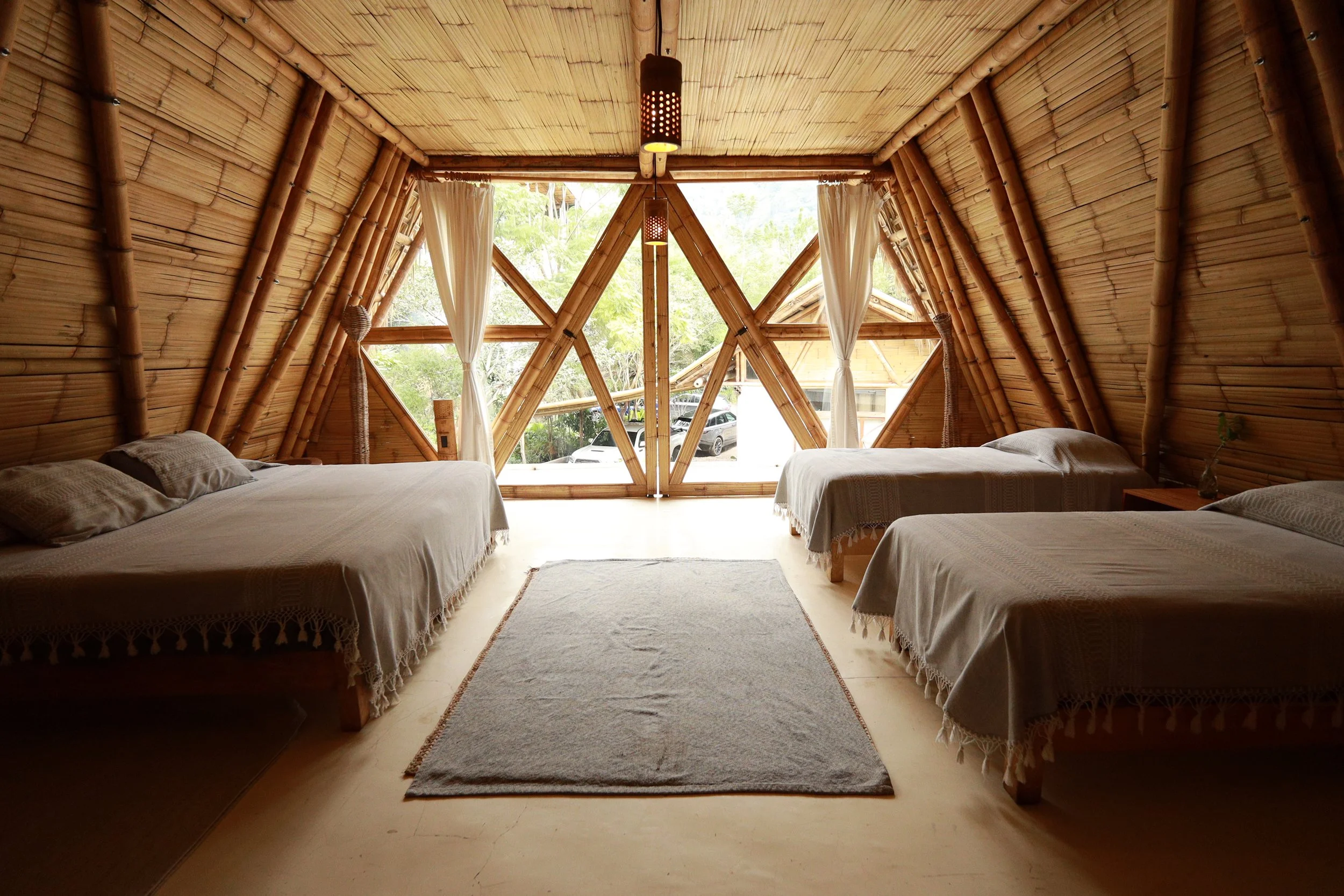 Dormitorio de madera con tres camas, cortinas blancas y vista a la naturaleza.