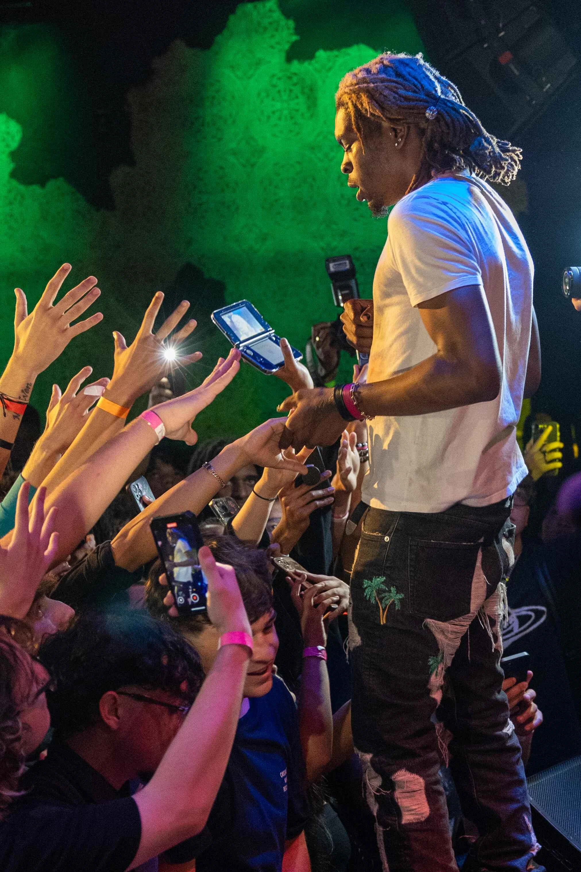 Performer with dreadlocks on stage during concert engaging with fans who are reaching out with smartphones.