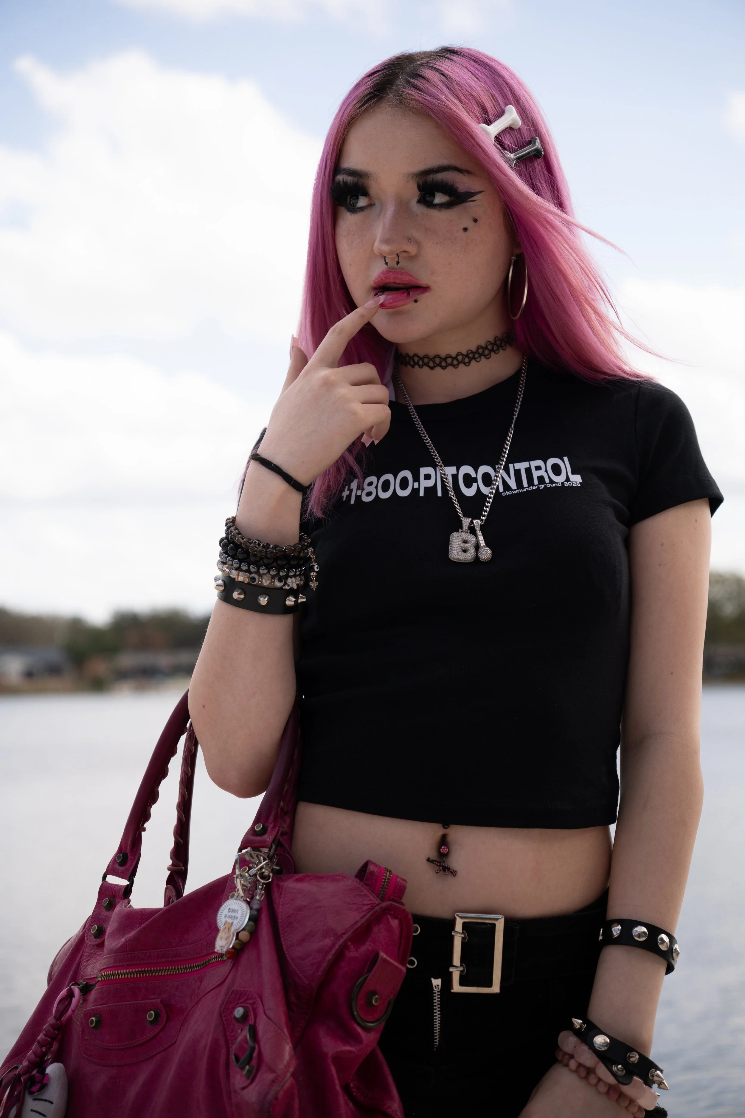Young woman with pink hair and piercings standing outdoors near water, wearing a black crop top with graffiti-style text, black pants with a metal belt, and multiple bracelets, carrying a pink handbag.