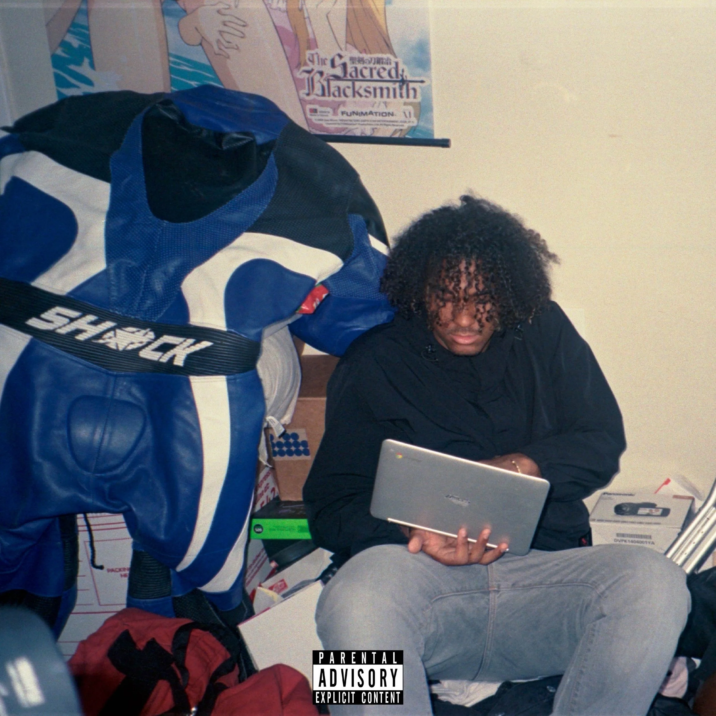 A person with curly hair sitting down and looking at a laptop, while another person in motorcycle gear stands next to them. There are posters and boxes in the background.