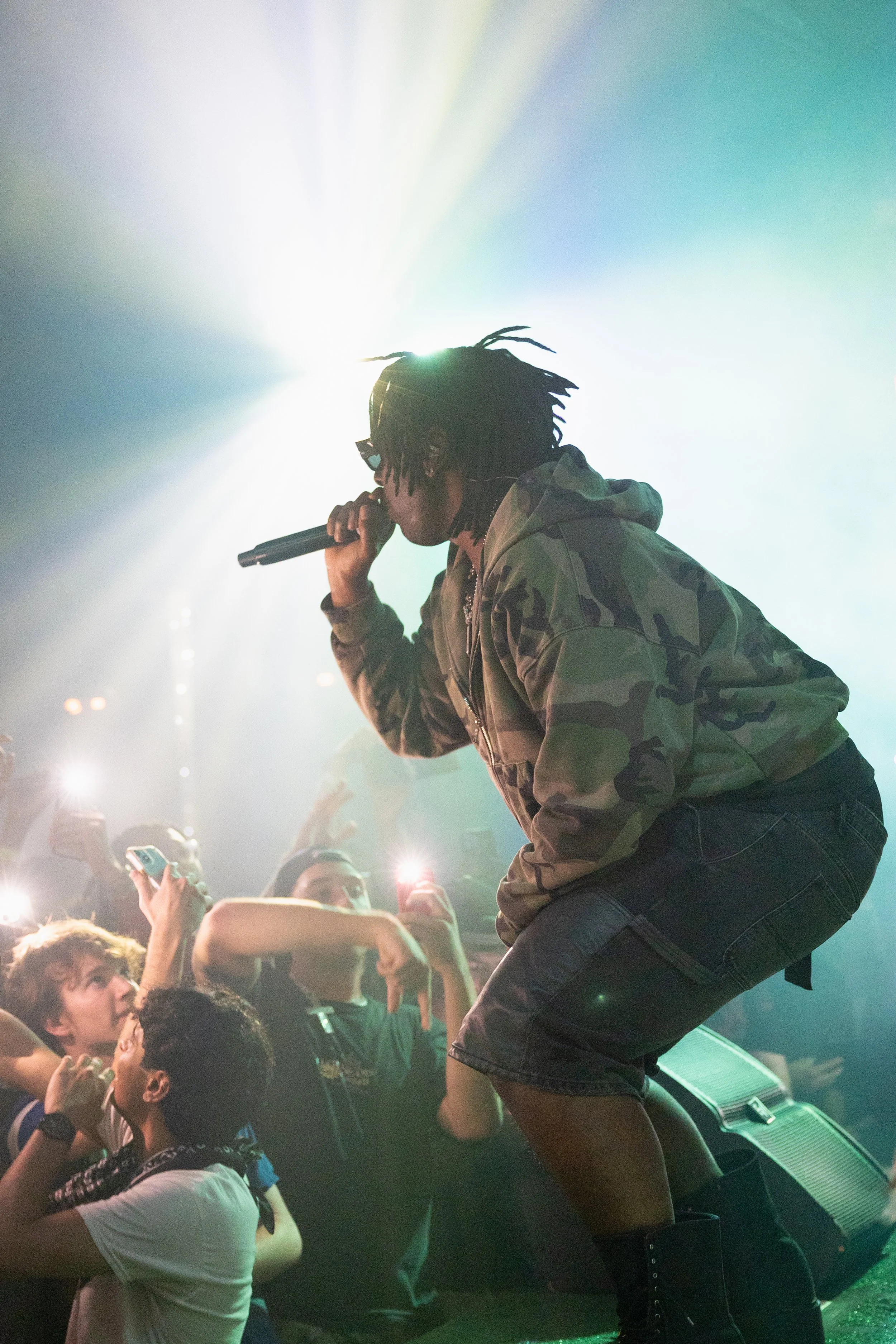 Performer singing into a microphone on stage, wearing a camouflage jacket and black shorts, with an audience taking photos and videos around them.