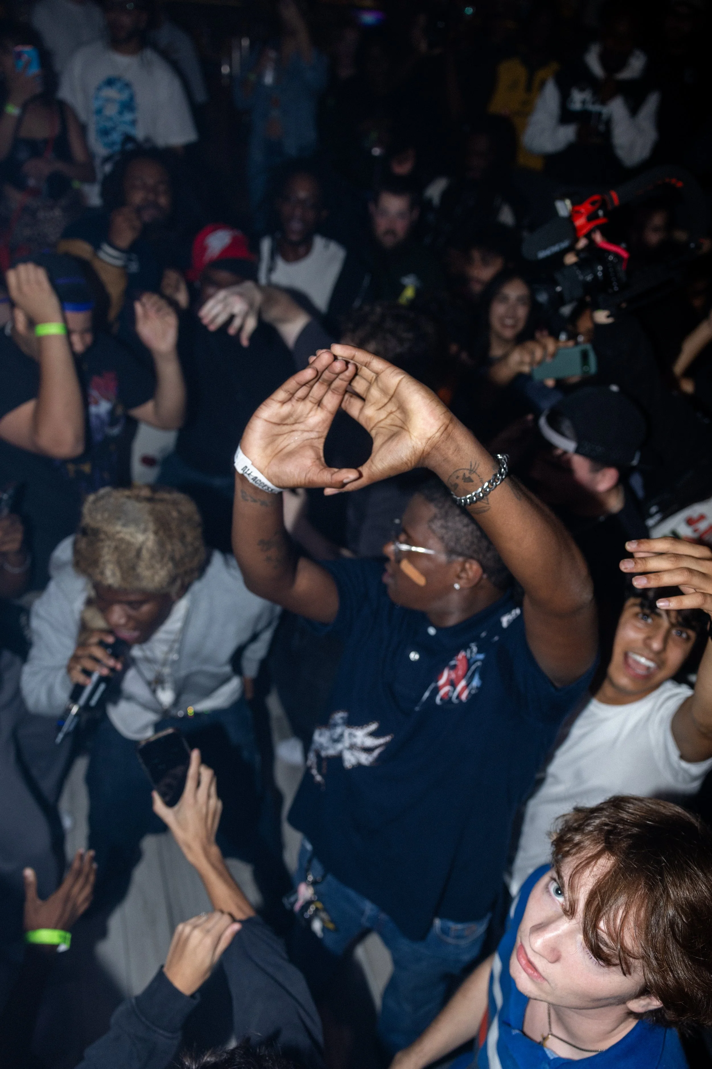 Crowd of people at a lively concert or event, with one person, wearing glasses and a dark shirt, making a hand sign and others smiling and holding microphones or phones.