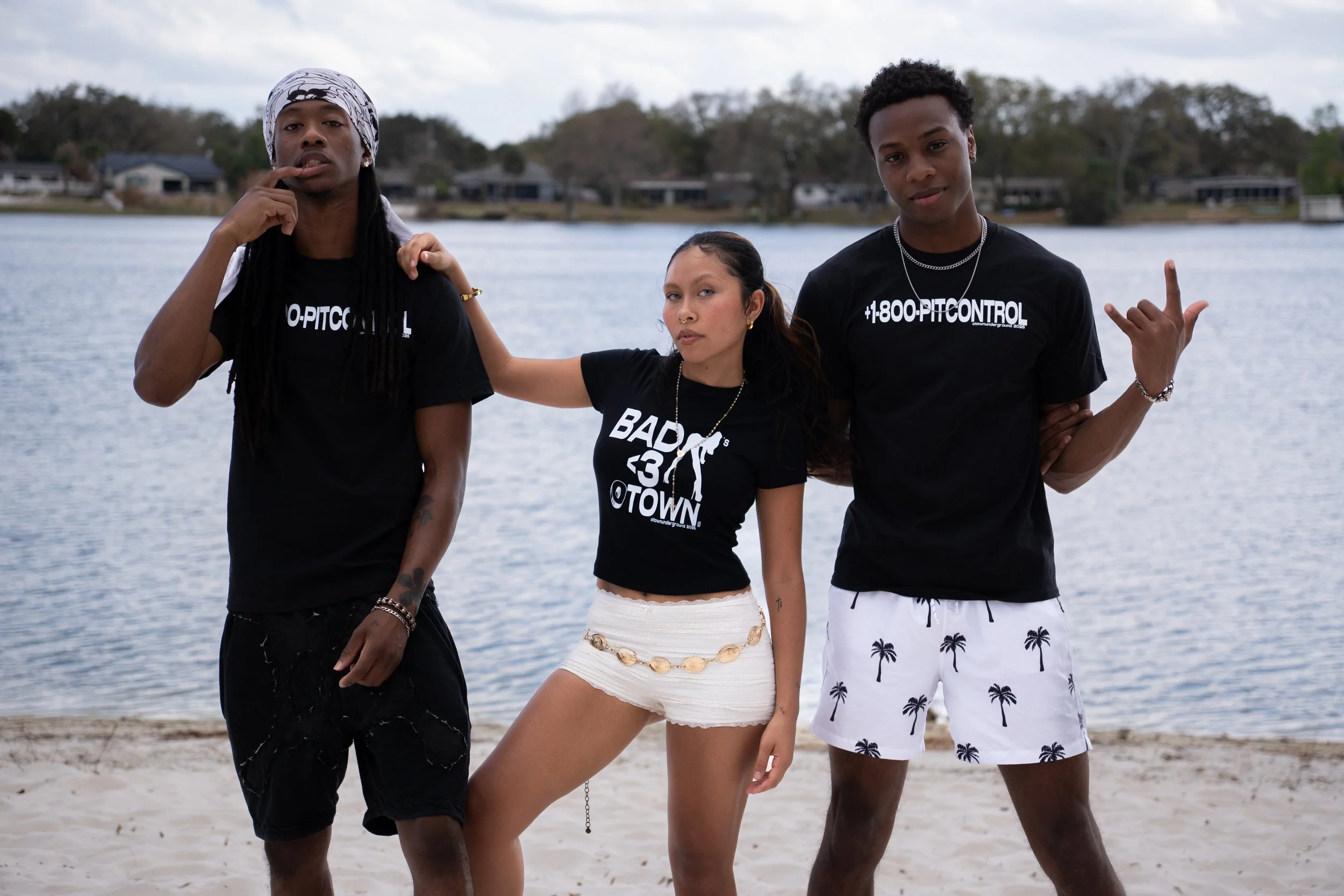 Three young people standing on a beach near a body of water, wearing casual summer clothing, with a cityscape and trees in the background.