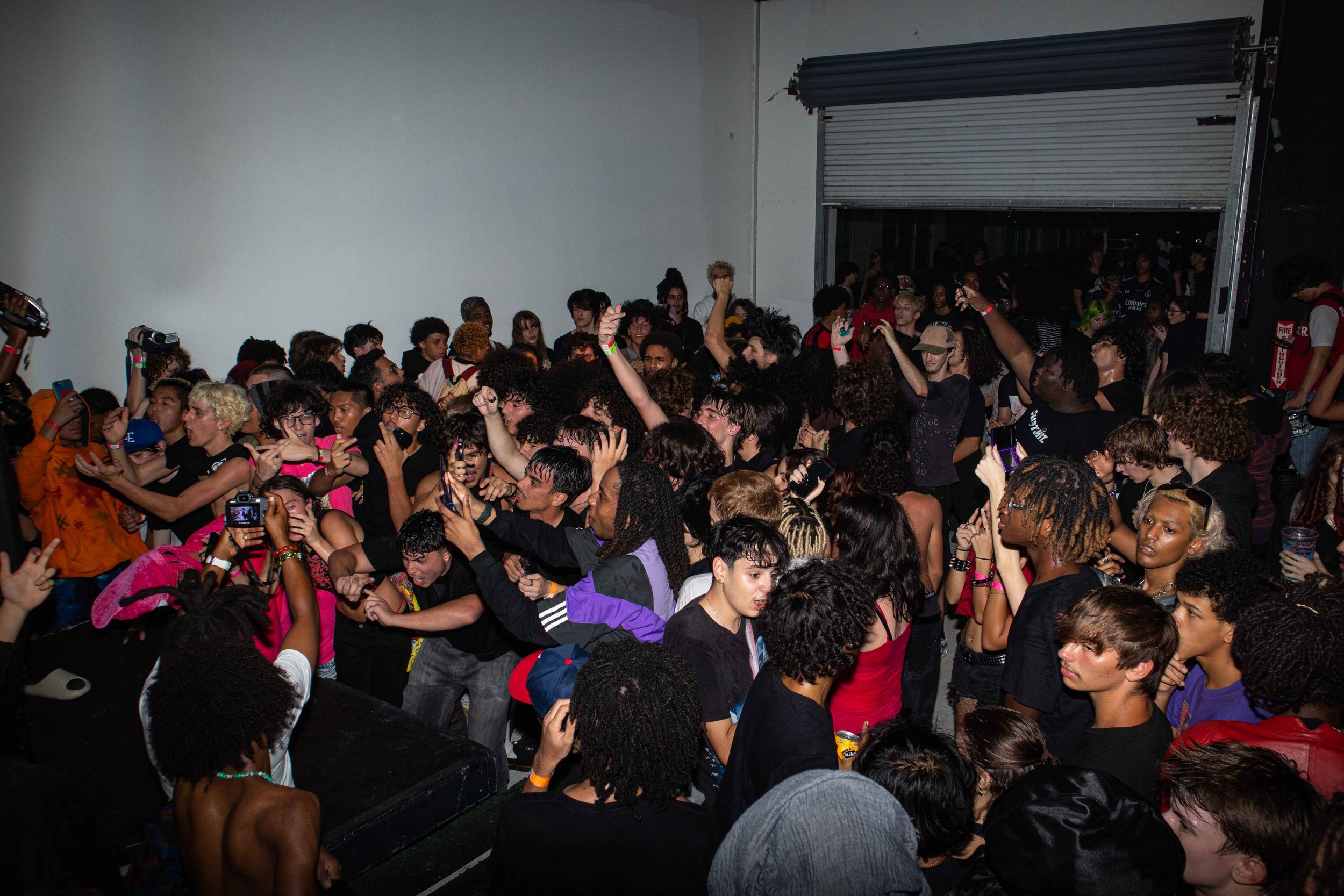 Young people at a crowded indoor concert or party, dancing and taking photos with their phones, with some wearing headphones and colorful outfits.
