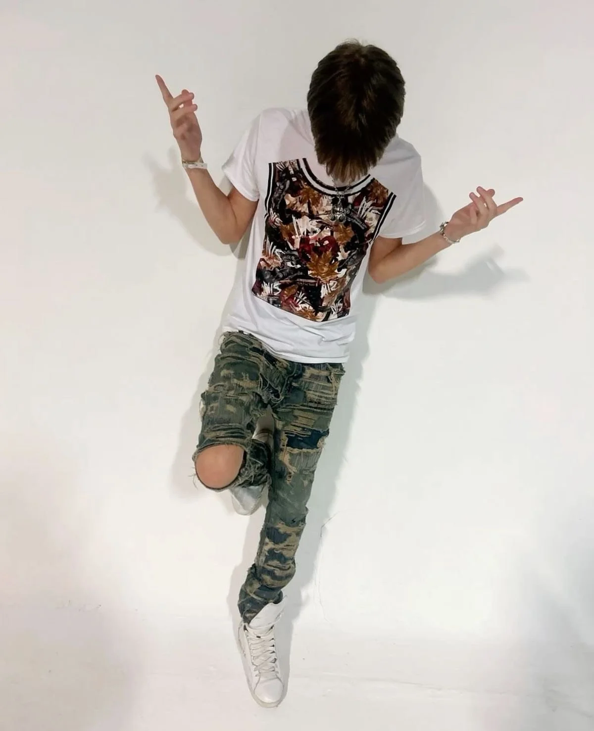 Person with brown hair, wearing a white graphic t-shirt, camouflage ripped jeans, and white sneakers, standing against a white wall with arms raised and head bowed.