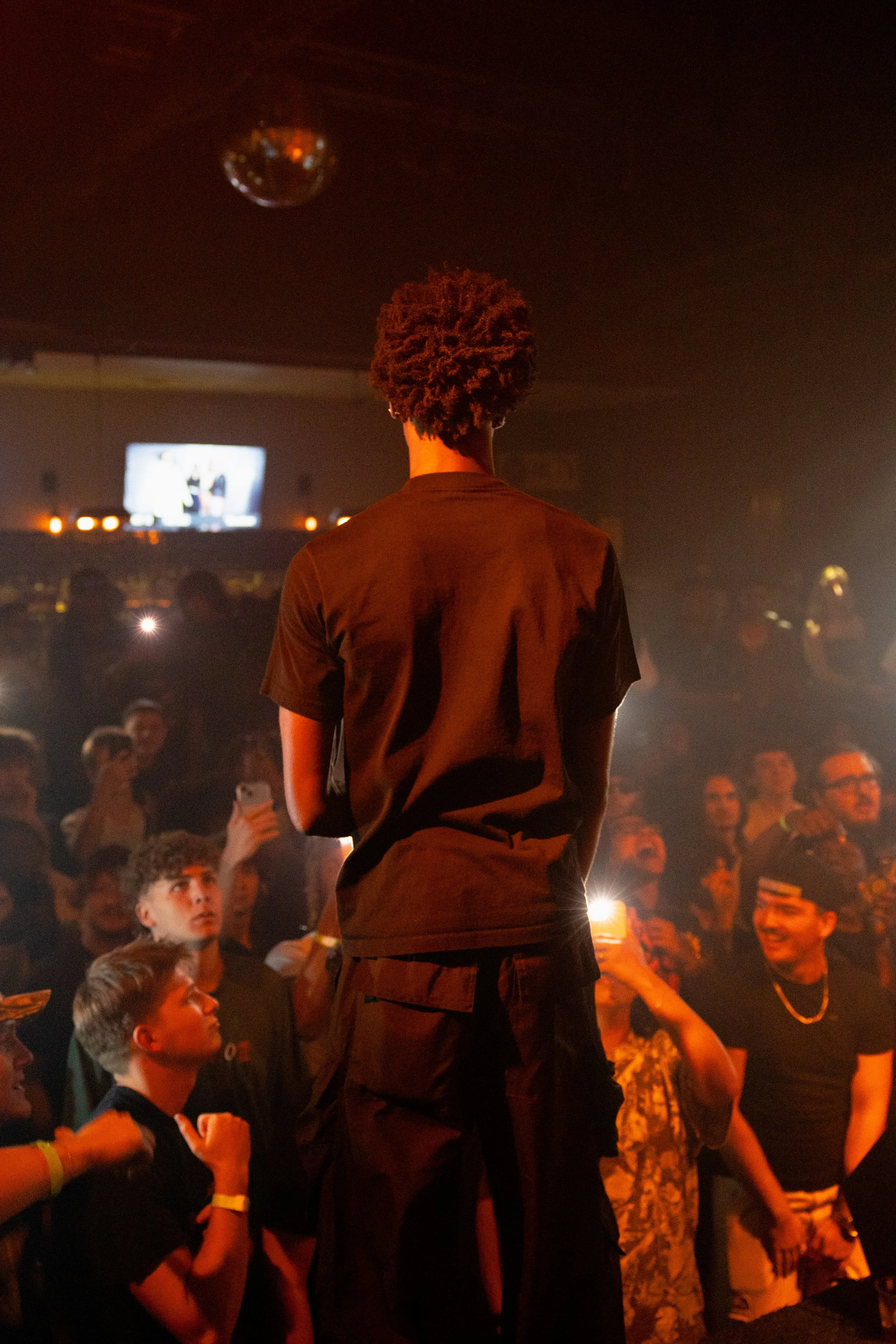 Person standing on stage facing an audience in a dimly lit venue with a disco ball hanging from the ceiling.