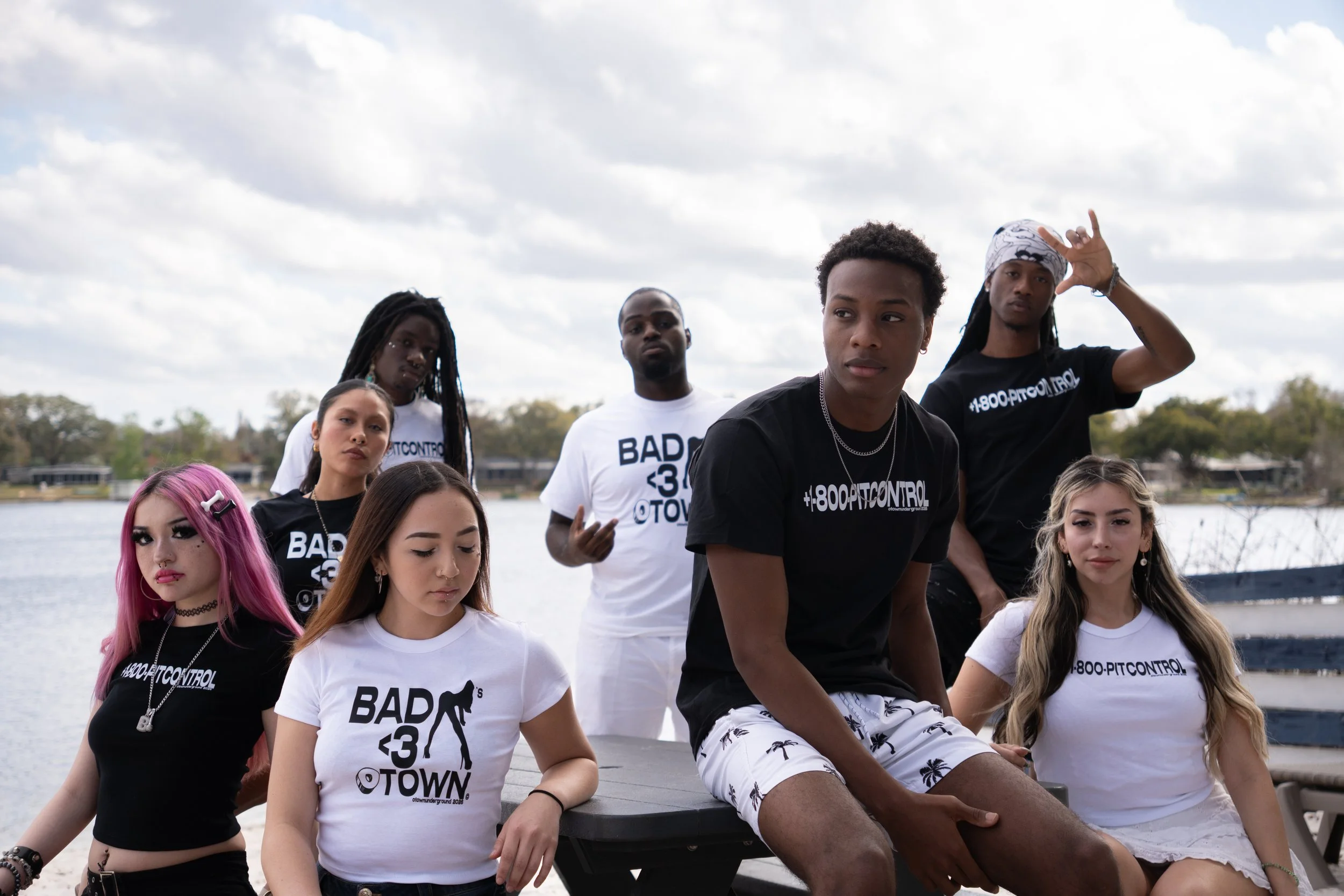 Group of young people sitting and standing outdoors near a body of water with trees in the background, wearing casual clothing and some with graphic t-shirts, under a partly cloudy sky.
