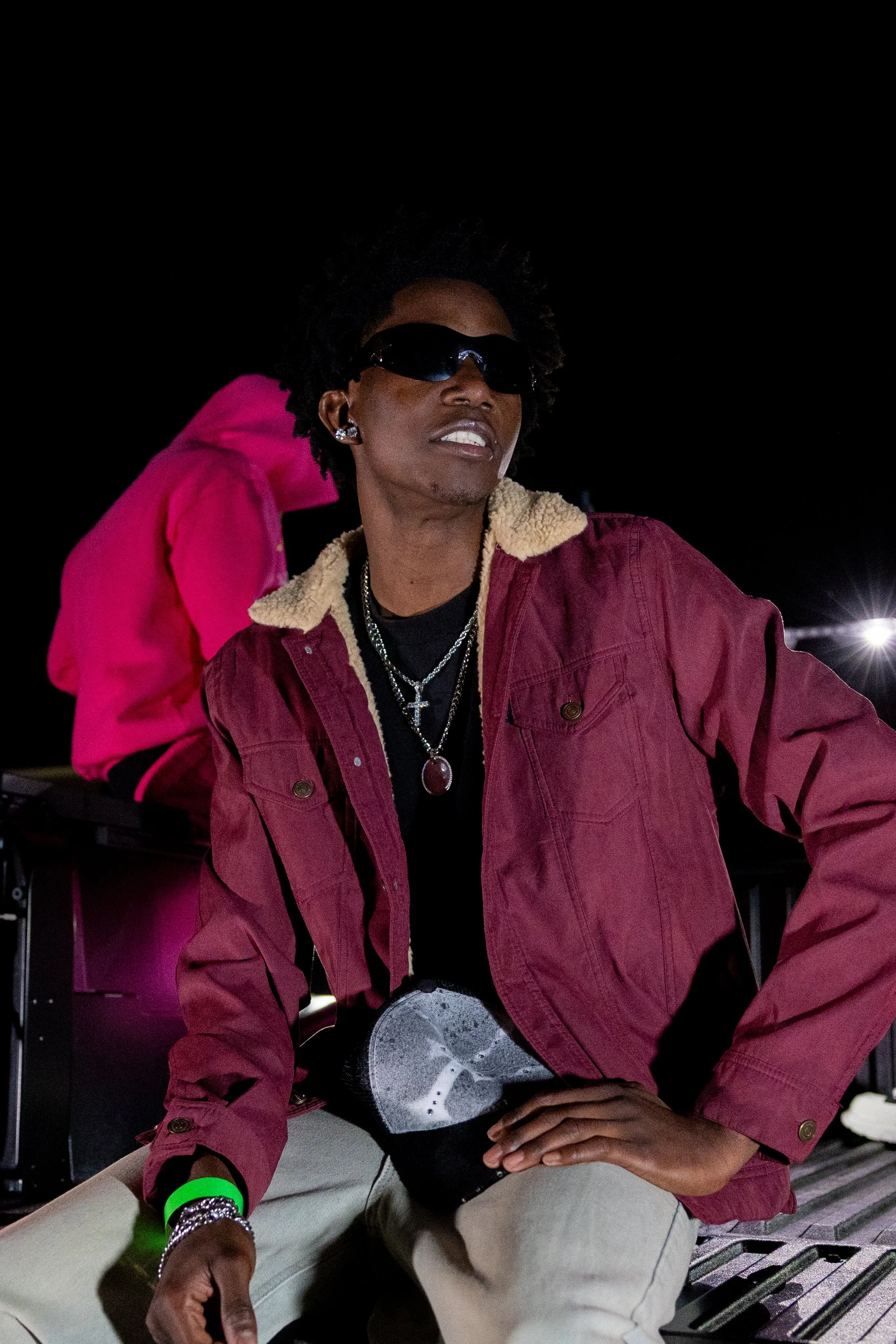 A man in a red jacket with beige lining, black shirt, and sunglasses sitting outdoors at night, with a woman in a pink hoodie and black pants behind him.