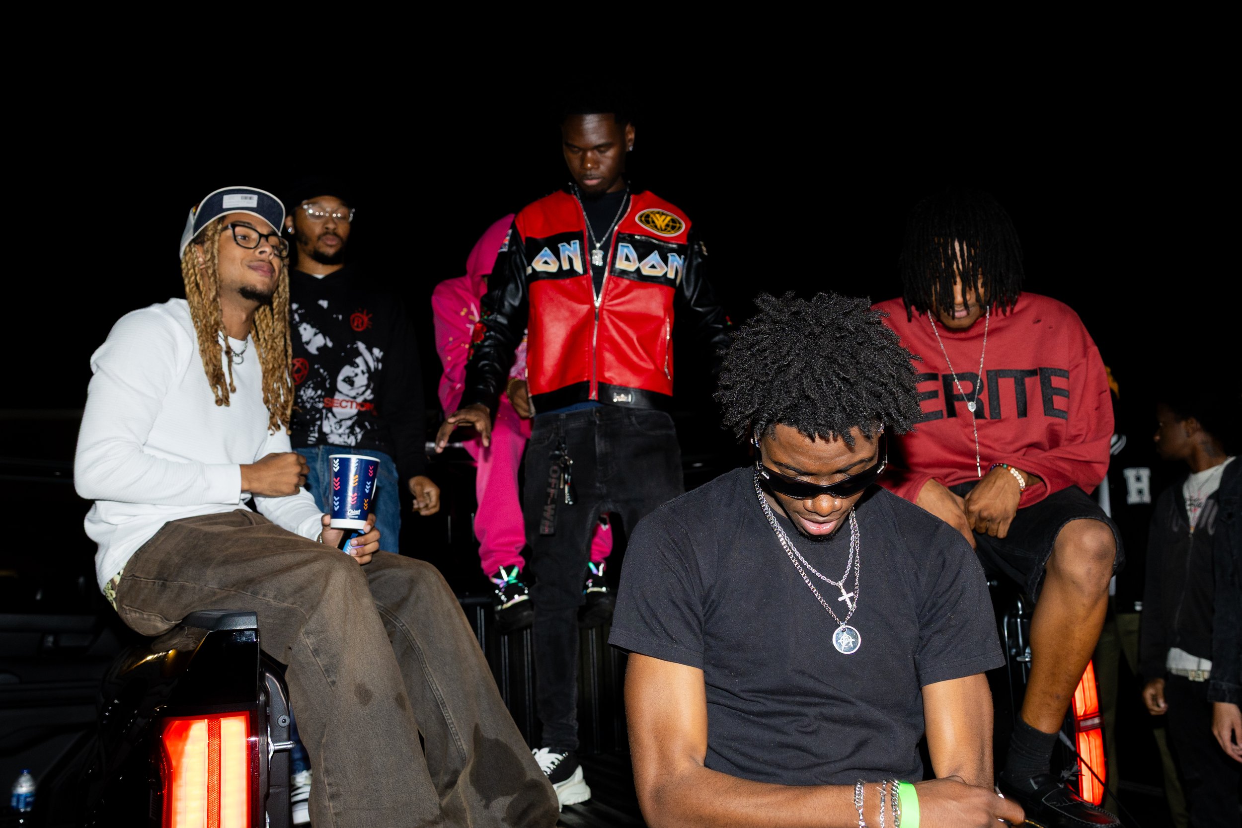 Group of young men at night, sitting and standing on a vehicle, dressed in casual streetwear, some with jewelry and drinks, in a social outdoor setting.