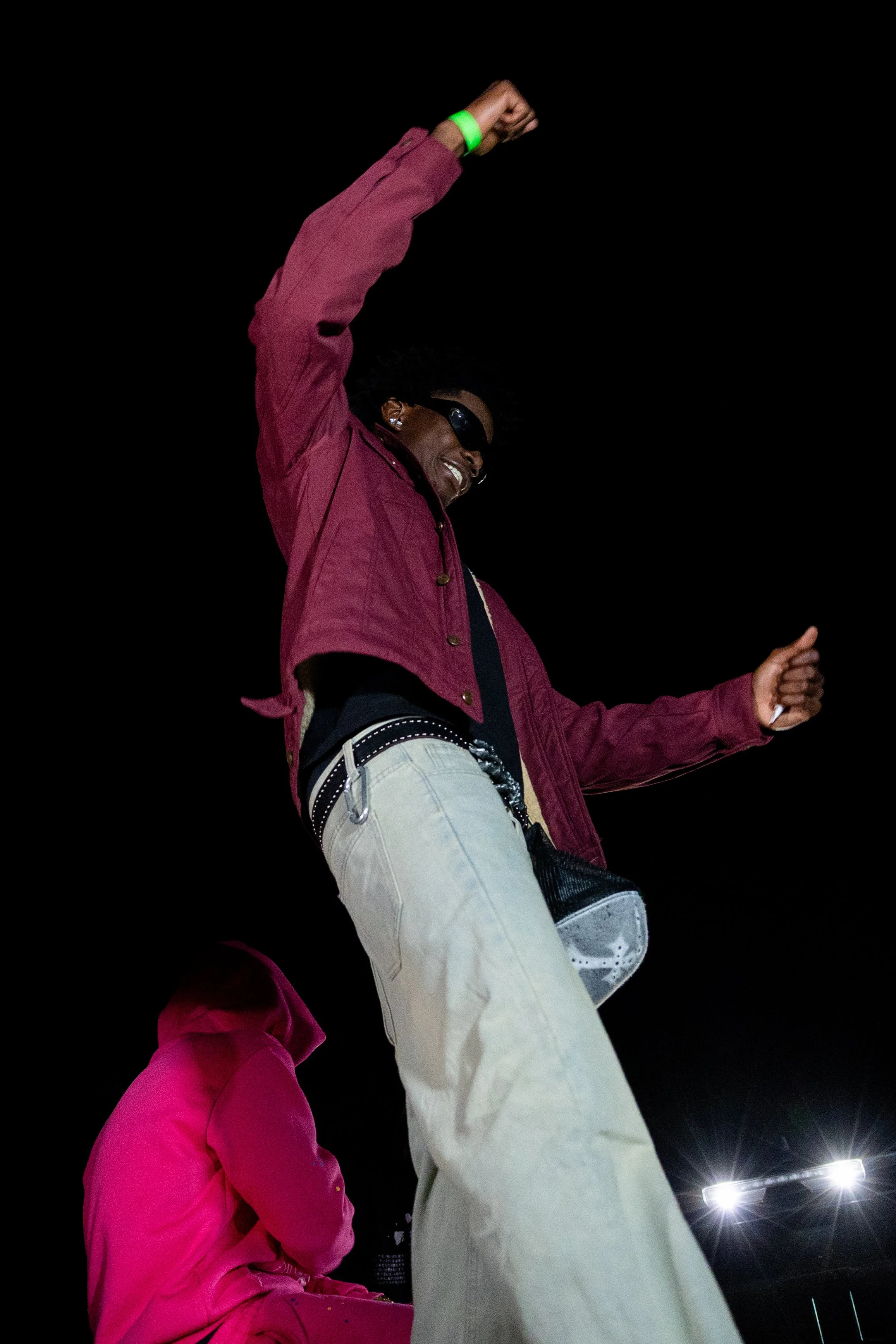 A person dancing outdoors at night, wearing sunglasses, a red jacket, and beige pants, with a dark sky in the background.