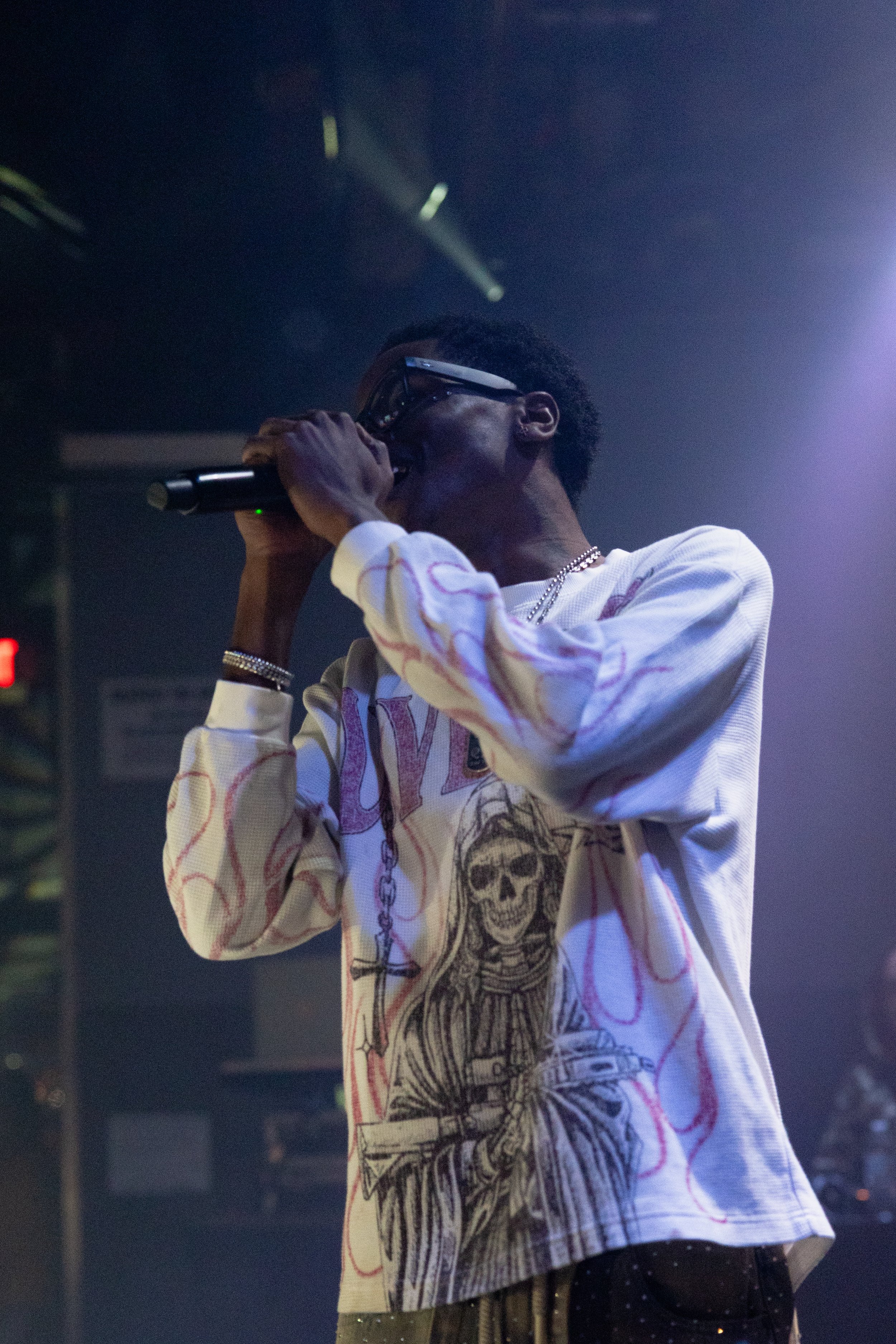 Person wearing glasses and a graphic white sweatshirt singing or speaking into a microphone on stage, with stage lighting and equipment in the background.