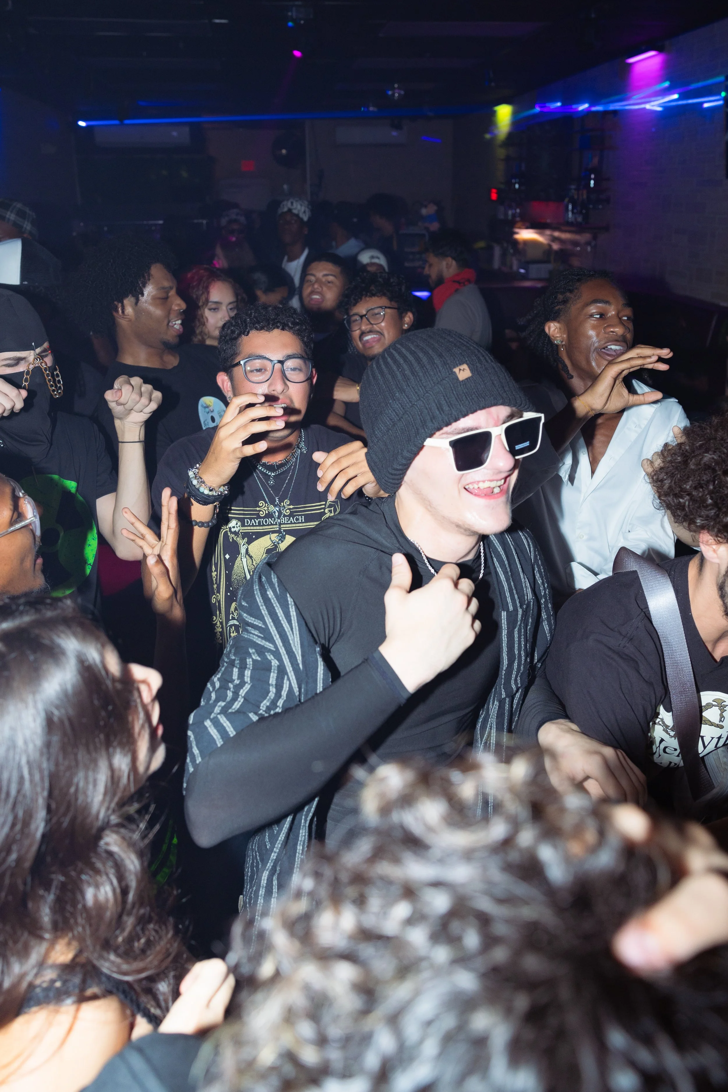 People dancing and enjoying themselves at a nightclub, with colorful lights and a bar in the background.