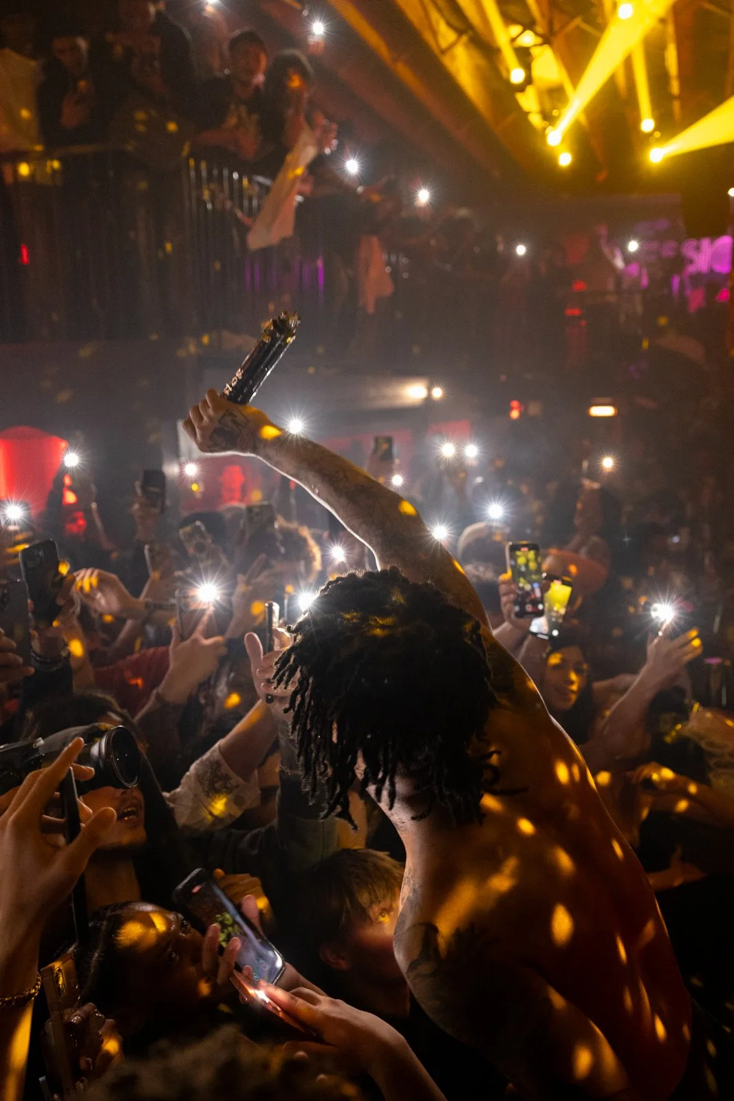 A person with dreadlocks holding a microphone crowd surfing at a concert or event, with numerous people taking photos and videos, bright lights, and a lively atmosphere.