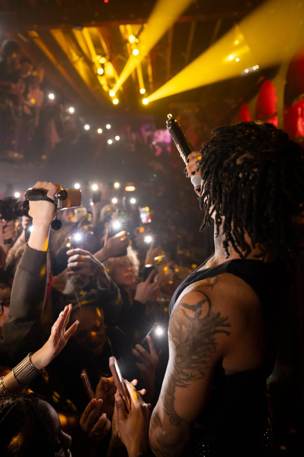 A performer with dreadlocks and tattoos on their arm is holding a microphone and engaging with a crowd at a concert or club, illuminated by yellow stage lights and surrounded by audience members taking photos with their phones.
