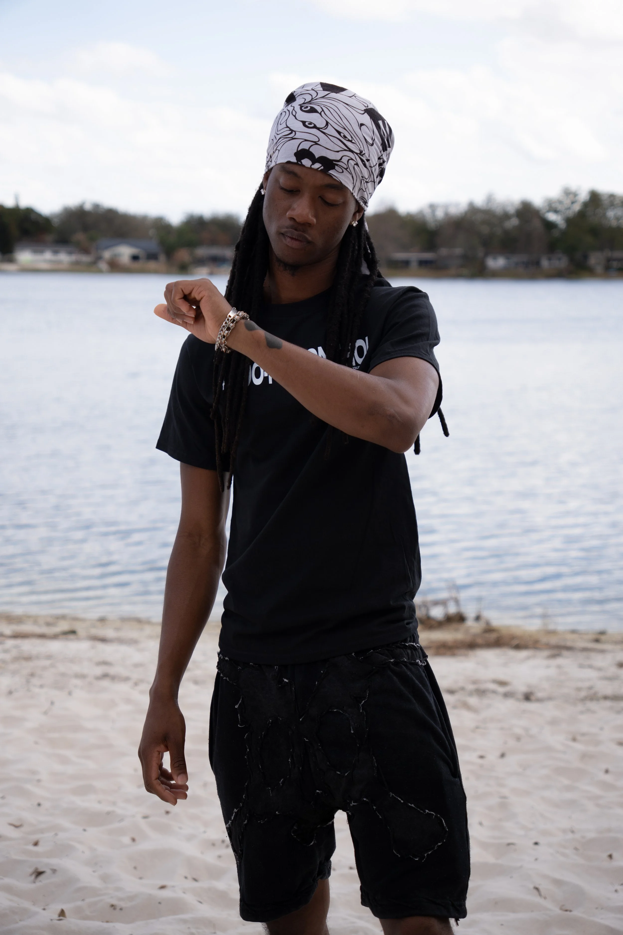 A young man with dreadlocks wearing a bandana, black t-shirt, and black shorts standing on a sandy beach by a body of water, looking at his wristwatch.