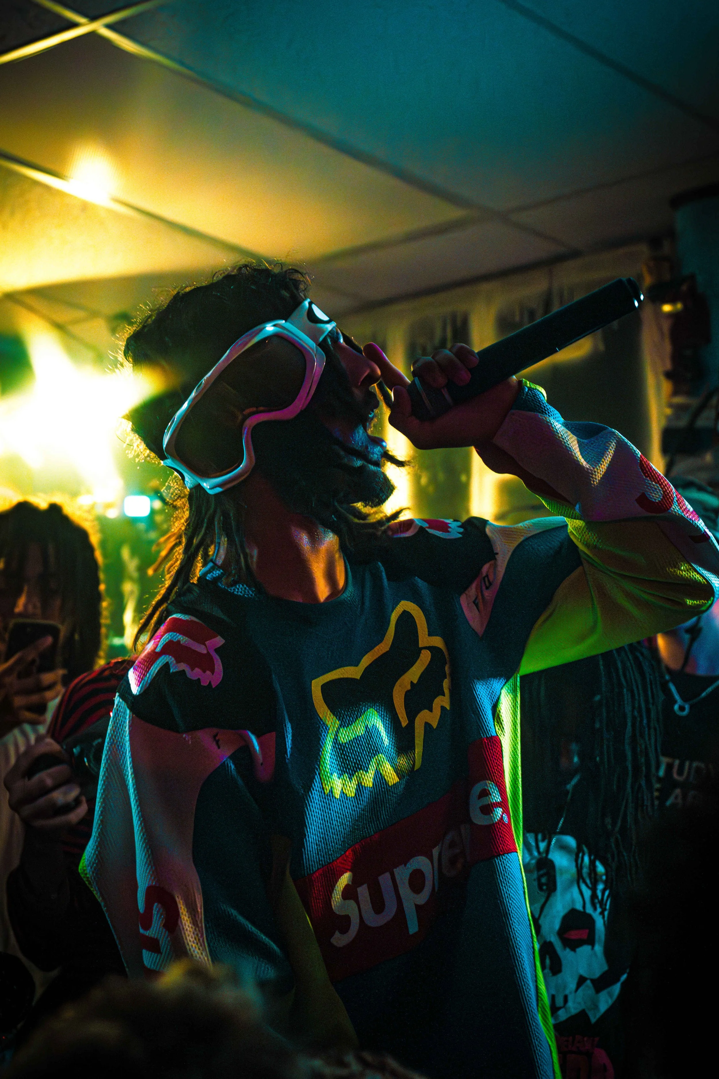 Person singing into a microphone at a party, wearing goggles and a colorful jersey with logos.
