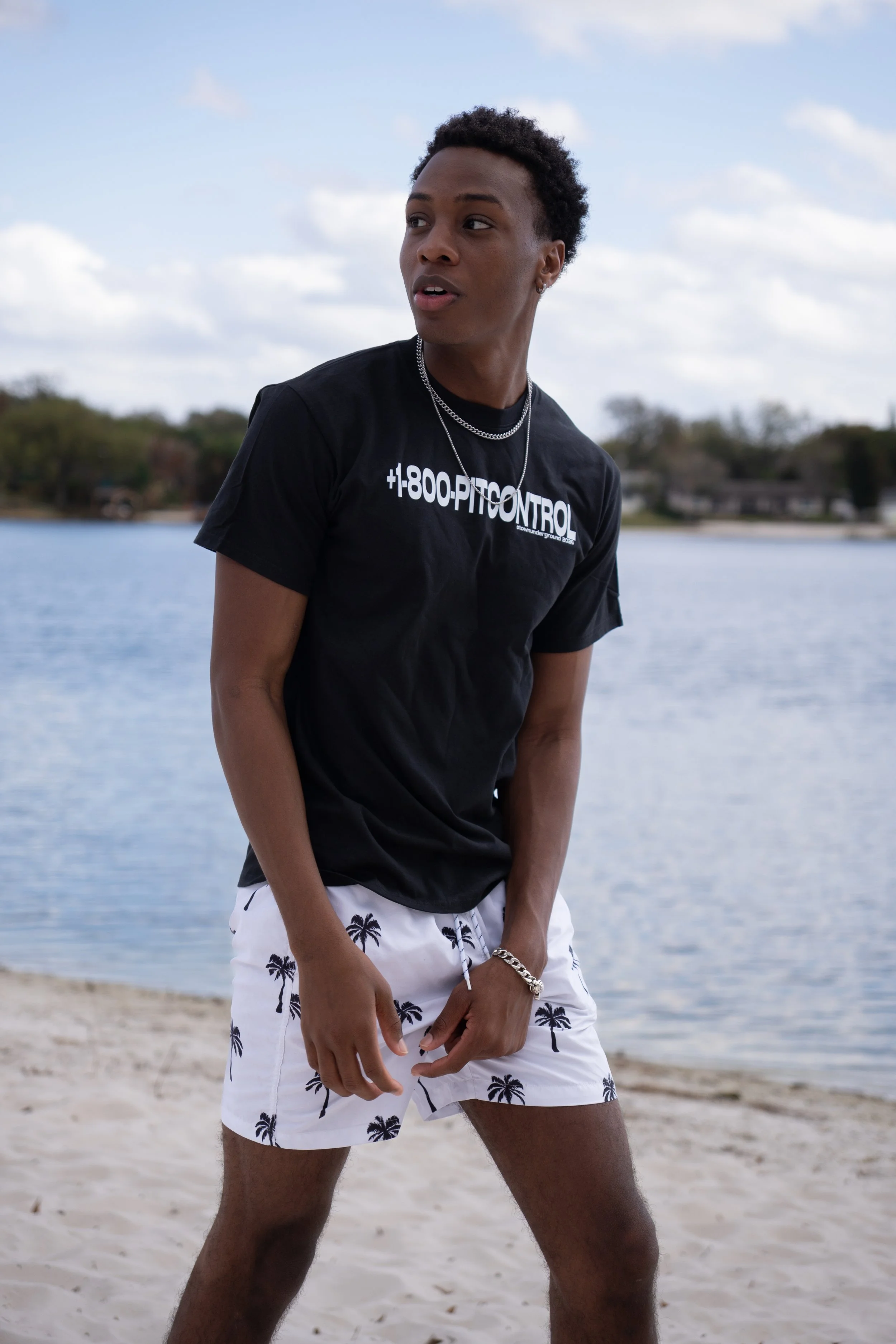 Young man standing on the beach near a lake or river, wearing a black T-shirt with white text and white shorts with palm tree patterns, looking to his right.