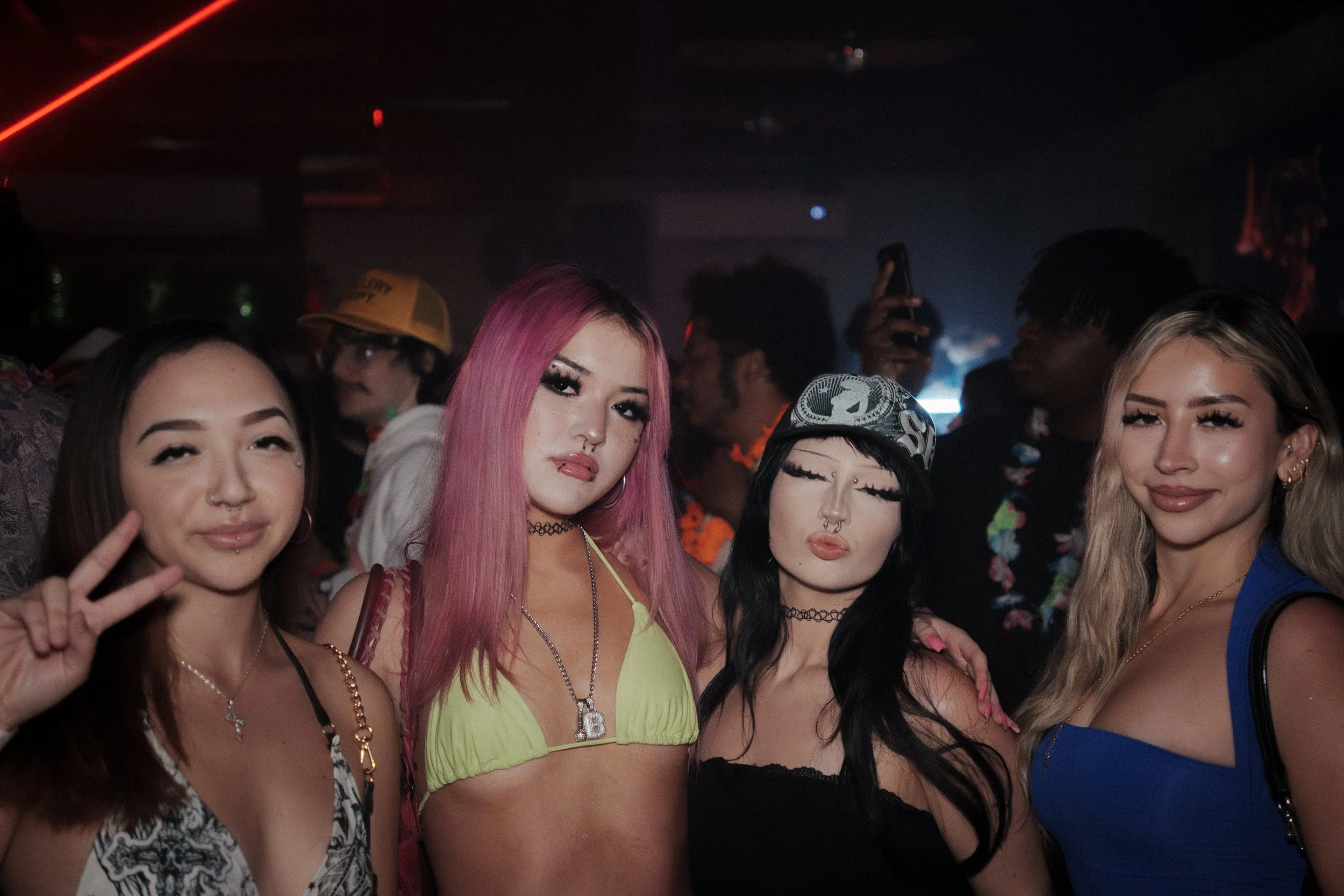 Four young women posing together at a nightclub, with a crowd and colorful lighting in the background.
