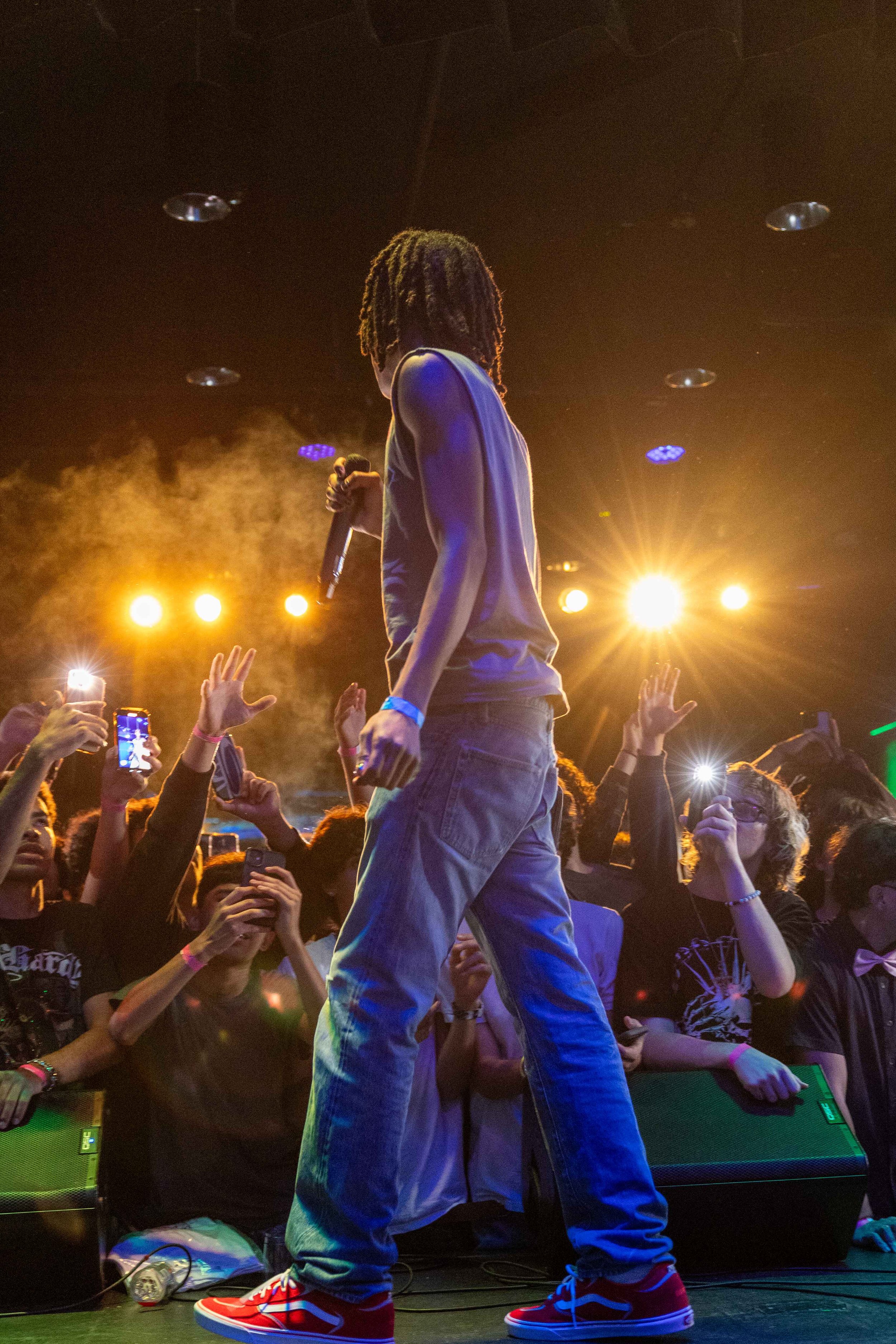 Performer with dreadlocks on stage holding a microphone, facing an audience with raised hands, bright stage lights illuminating the scene.