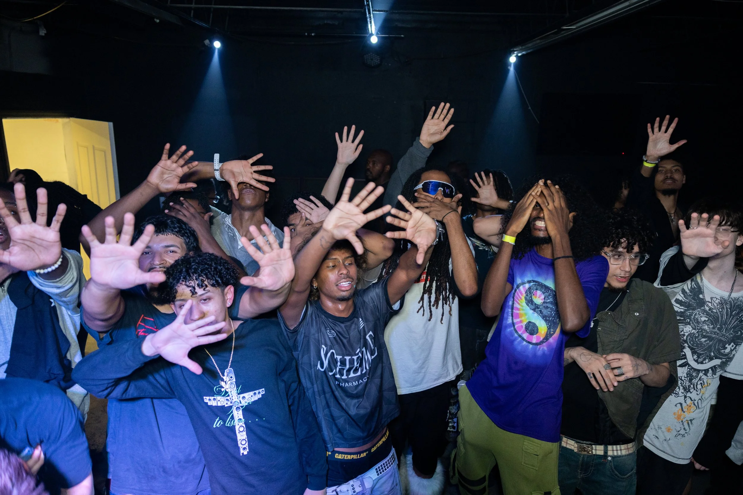 Group of people at a concert or event with many raising their hands, some covering their faces, in a dark indoor setting with blue lighting.