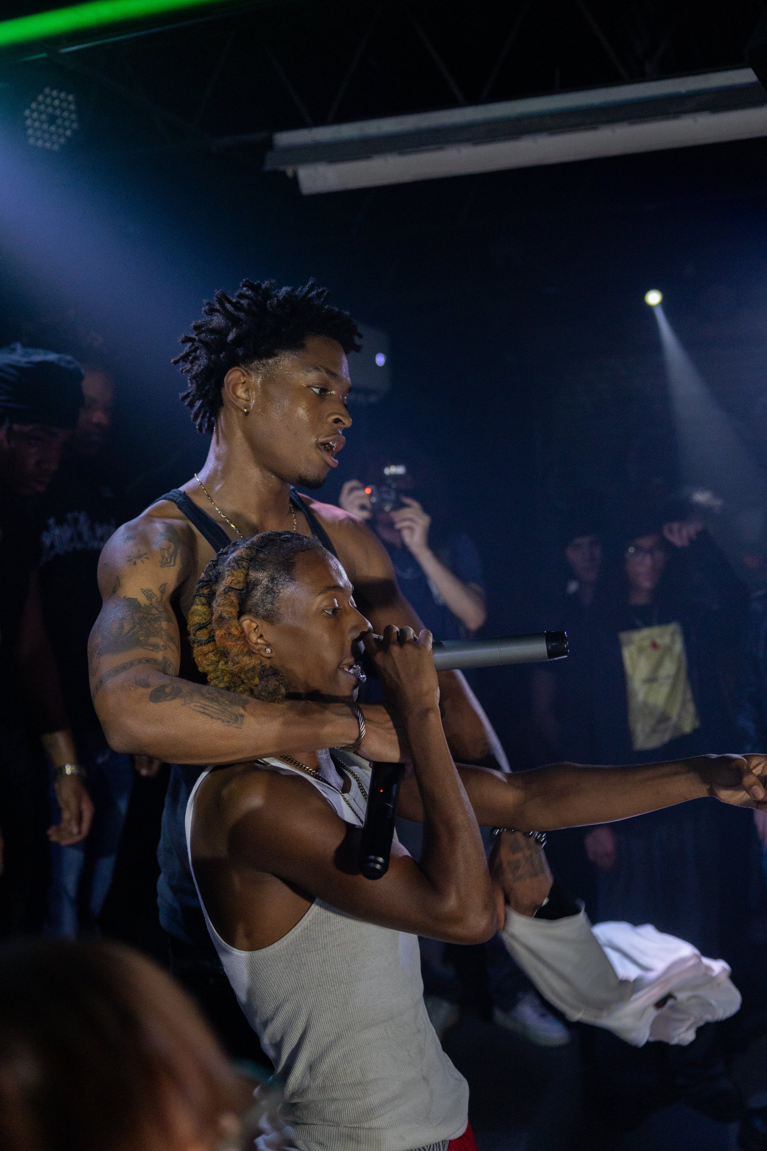 A young male performer with tattoos and dreadlocks embracing a young woman with braided hair, holding a microphone and singing or rapping on stage inside a dark club or concert venue.