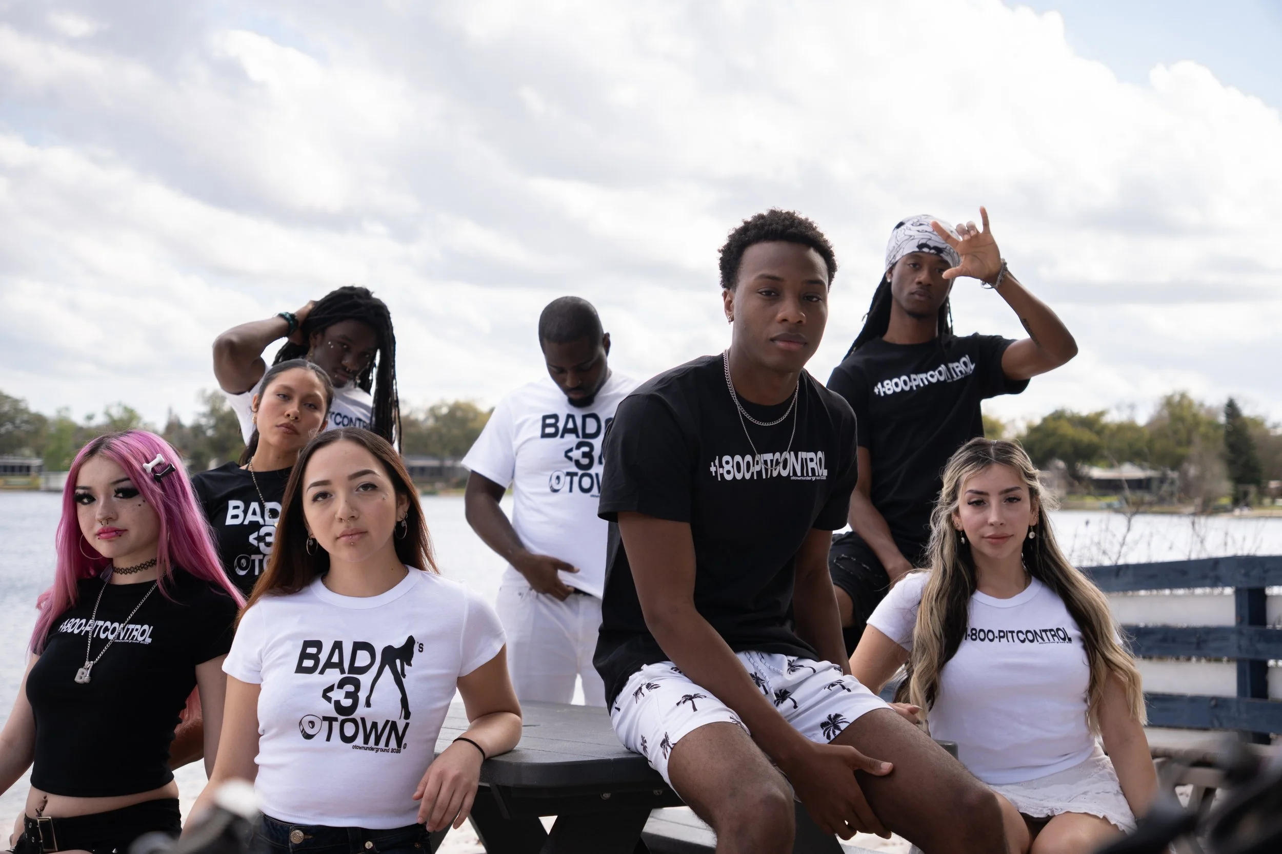Group of seven young people sitting and standing outdoors near water, wearing casual clothes with various graphic text, some with pink and blonde hair, some with dreadlocks, posing for a photo on a cloudy day.