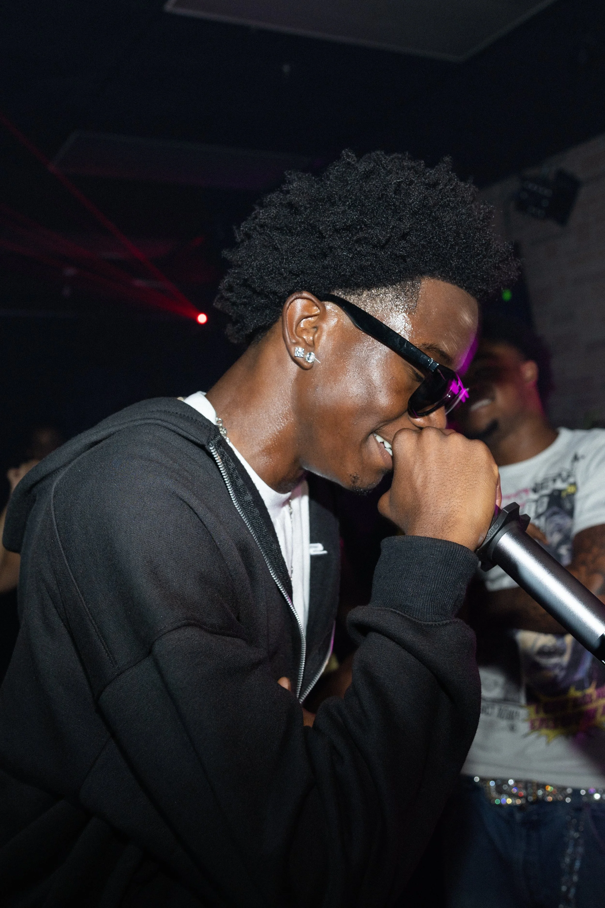 A young man with glasses and earrings singing or rapping into a microphone in a dimly lit club or party setting.