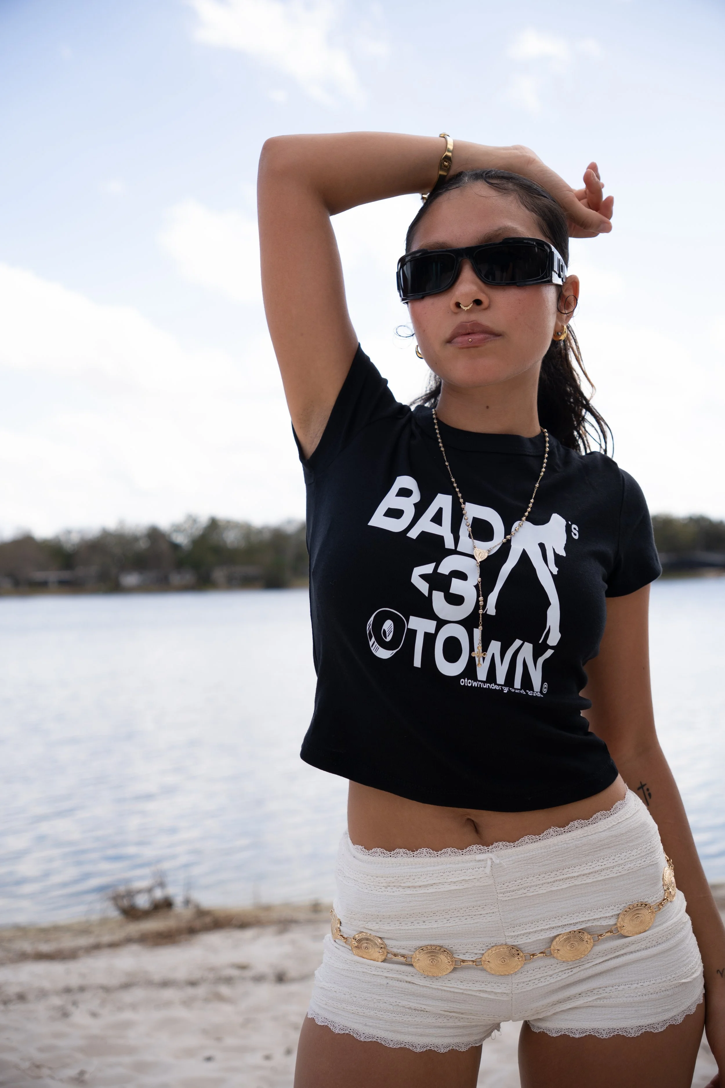 A woman with sunglasses and tattoos wearing a black t-shirt with text and a graphic, beige shorts with a decorative belt, standing outdoors near a body of water.