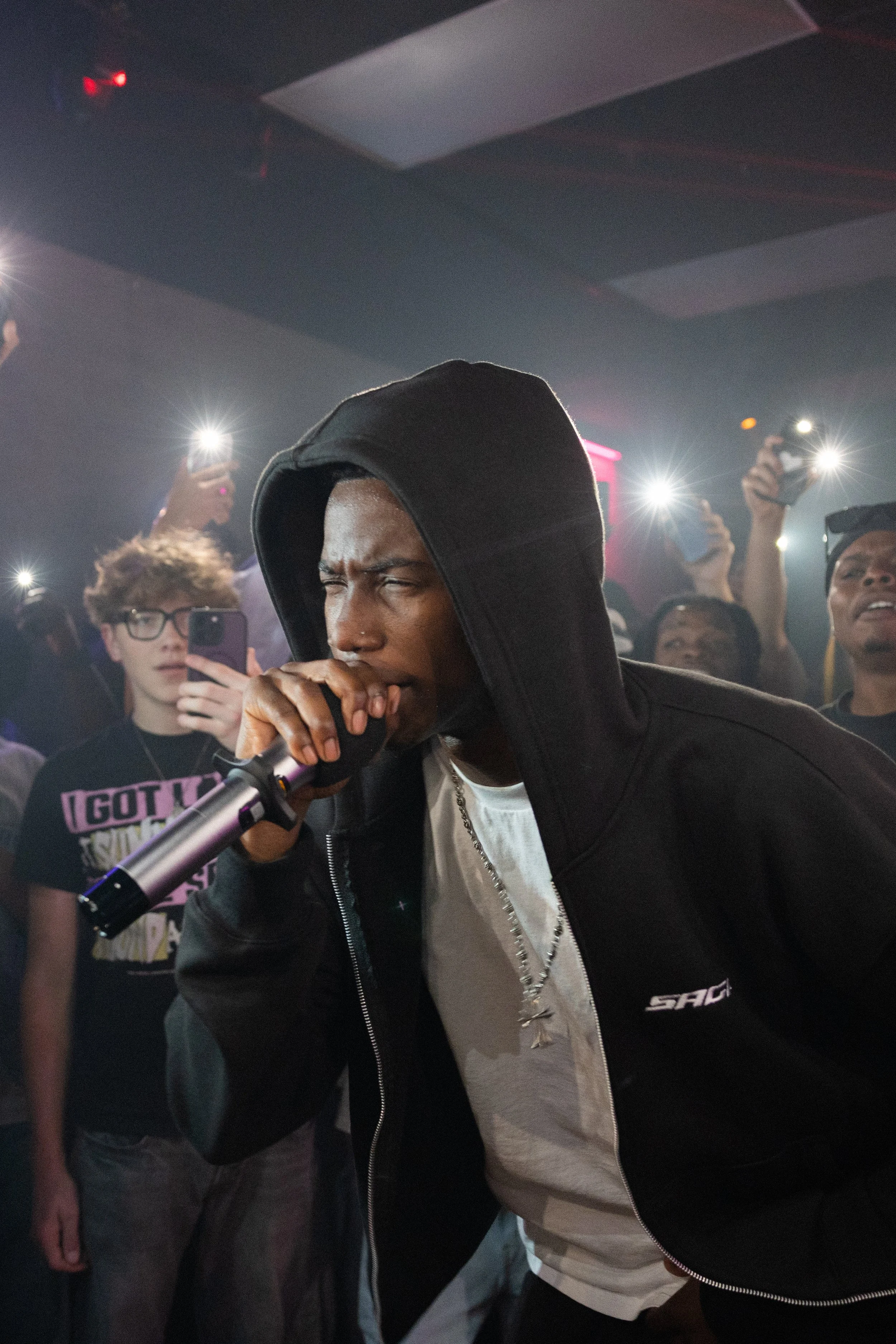 A male performer wearing a hoodie and a chain sings into a microphone on stage at a concert, surrounded by an audience capturing the moment with their phones.