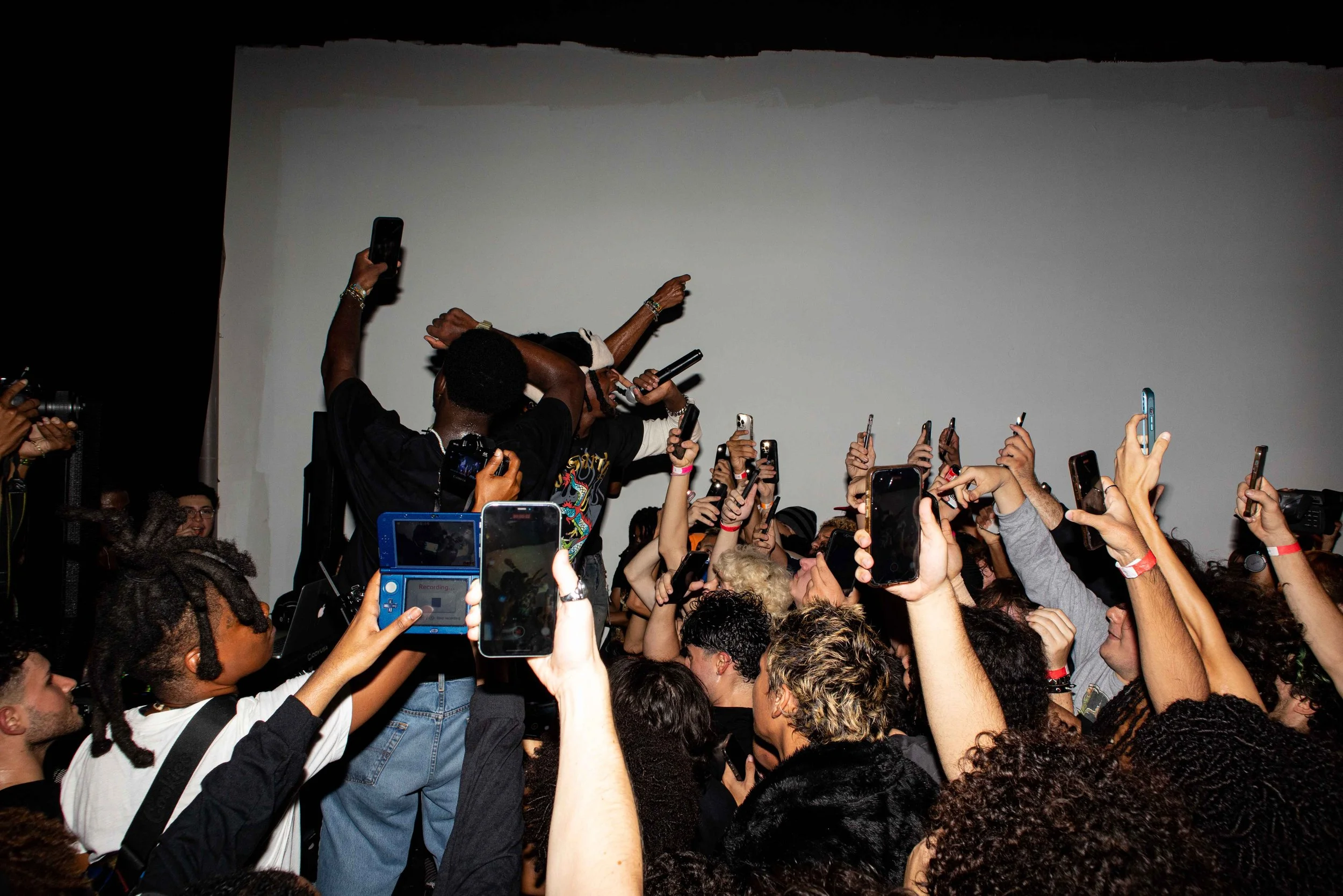 A crowded concert or party scene with many people holding up their phones to record or take pictures of a performer on stage who is holding a microphone.