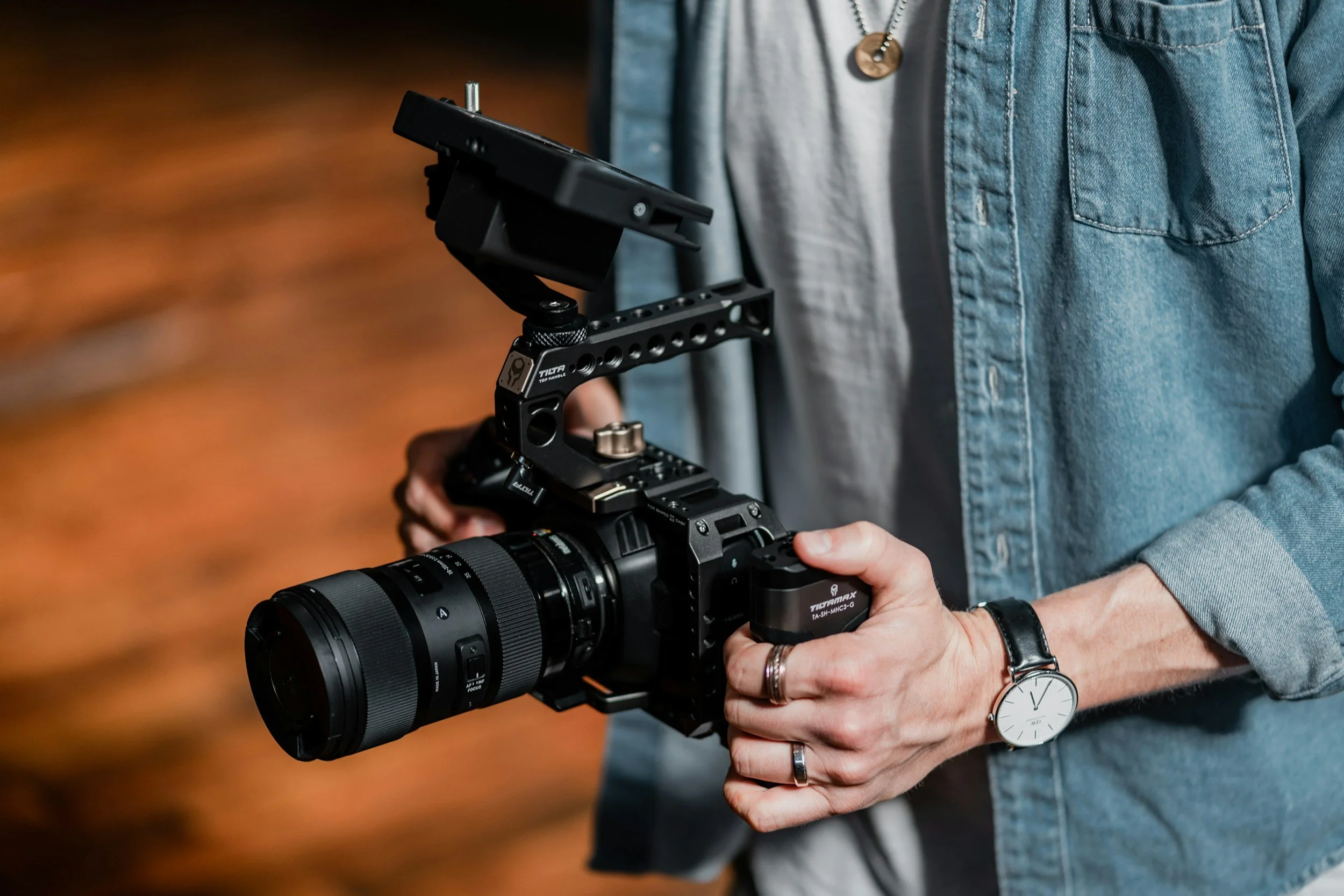 Person holding a professional camera with a mounted monitor in a wooden-floored room.