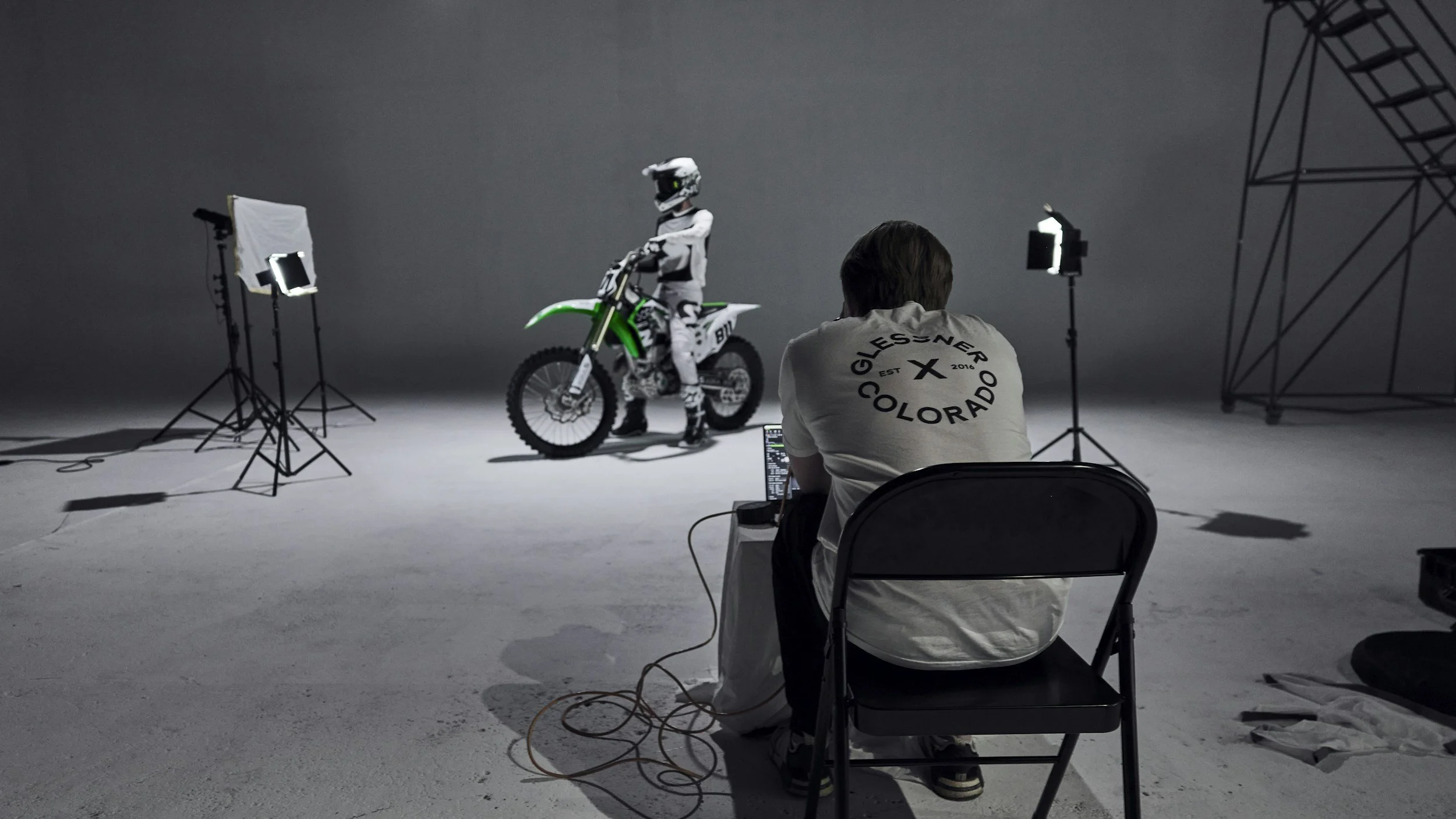 A photoshoot scene with a person sitting on a black folding chair, wearing a white shirt with black text, photographing a motocross rider on a green dirt bike. The rider is wearing a helmet and protective gear, standing in a studio with lighting equipment and a gray backdrop.