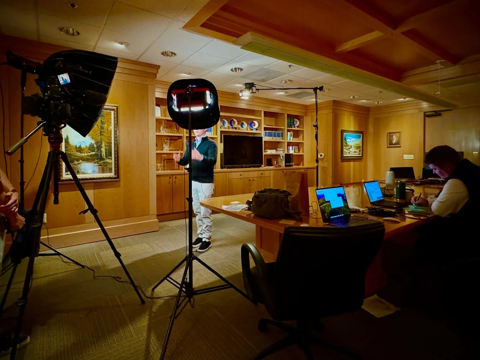 A professional video recording setup in a wood-paneled office room with a woman speaking to a camera, two laptops on a table, and a person taking notes.