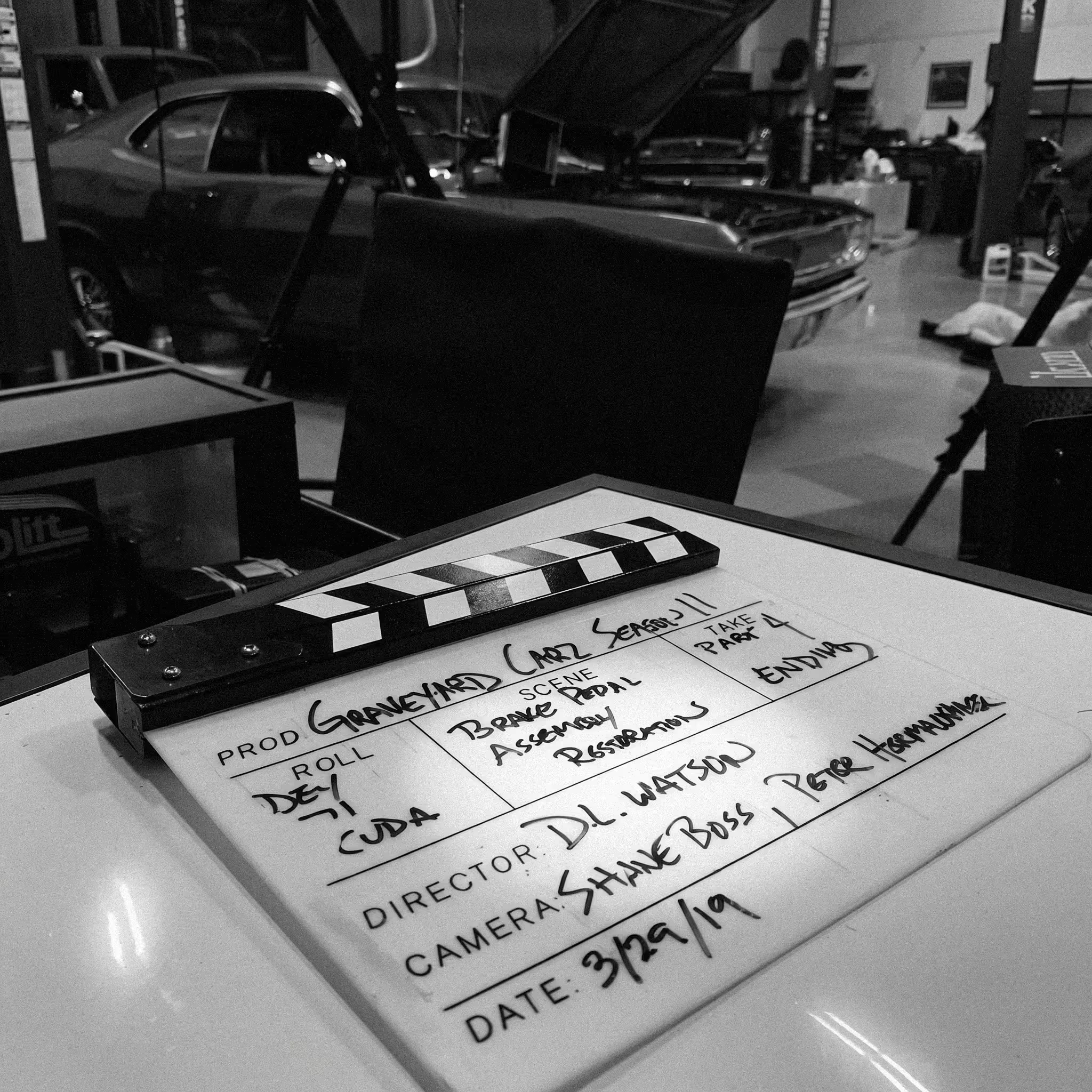 Black and white photo of a clapperboard with handwritten scene and take information, placed on a table near a model car and other objects in a room that resembles a workshop or storage area.