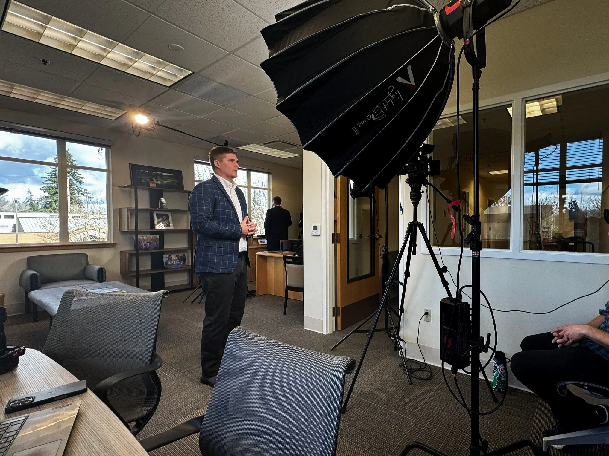 A man in a checked blazer and white shirt standing in front of a camera setup with a large umbrella light, inside an office or conference room with large windows, chairs, and tables.