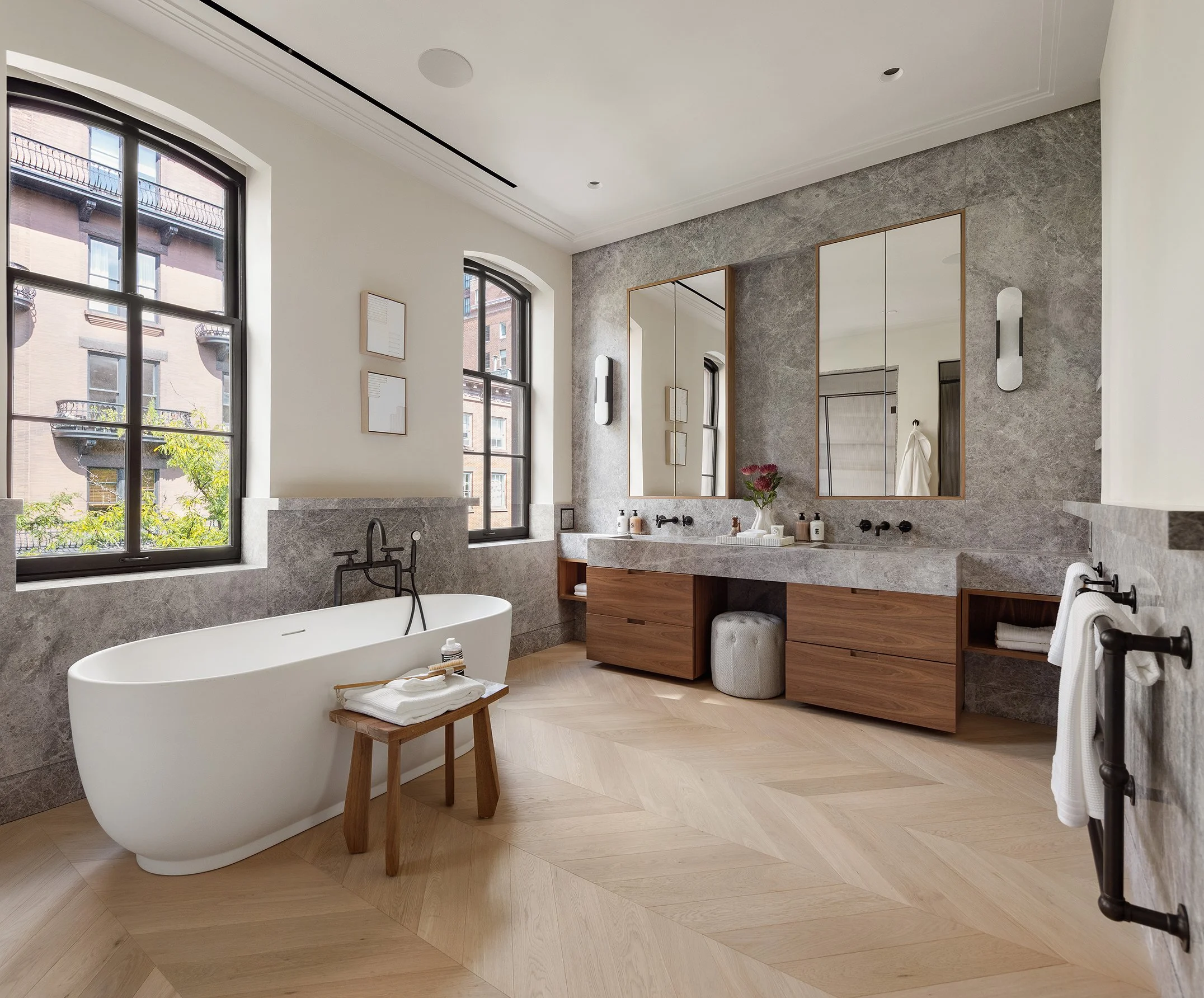 Modern bathroom with freestanding bathtub, double vanity with two mirrors, large windows, and light-colored wooden flooring.