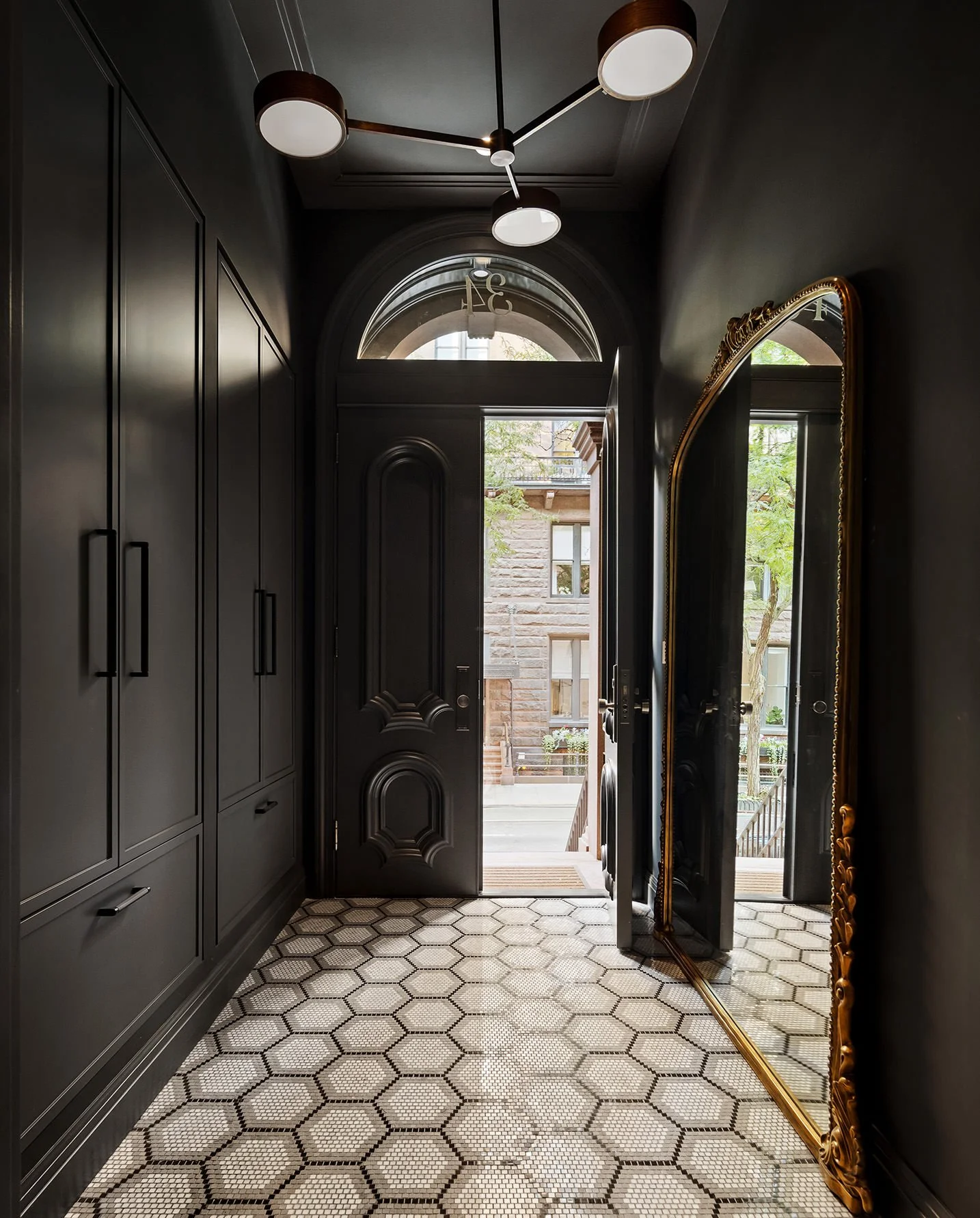 Entryway with black walls, hexagon-tile floor, large mirror with ornate gold frame, black front door, and a modern chandelier with four round lights.