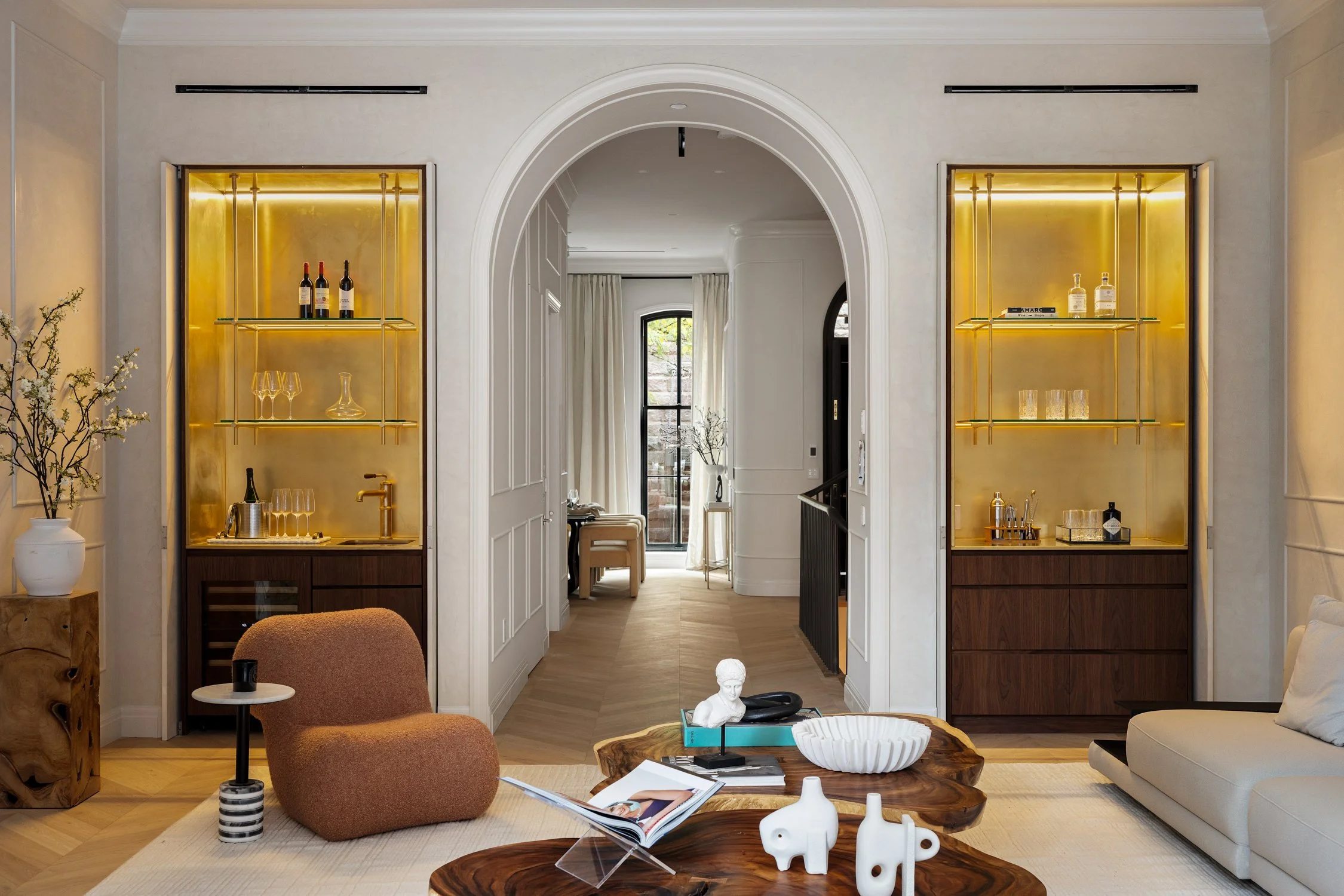 Living room with built-in glass shelves, gold backing, and bottles of wine, glassware, and bar accessories. A doorway leads to a room with a window, curtains, and seating area. Modern furniture and decorative items are arranged inside.