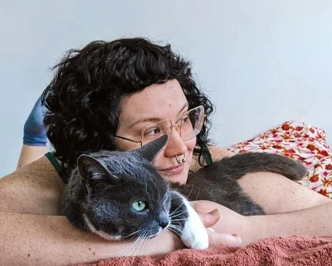Person with curly hair, glasses, and septum piercing lying on a bed with a black and white cat, both looking relaxed.