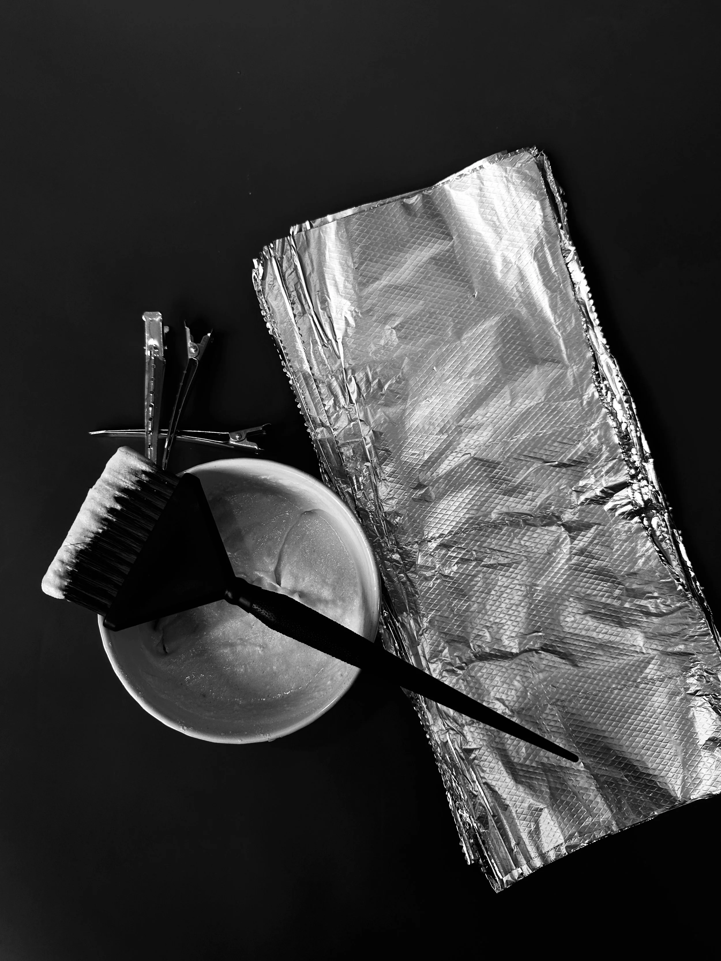 A baking setup with a cup and a silicone brush resting on a plate, foil-lined baking tray, and three skewers on a dark surface.