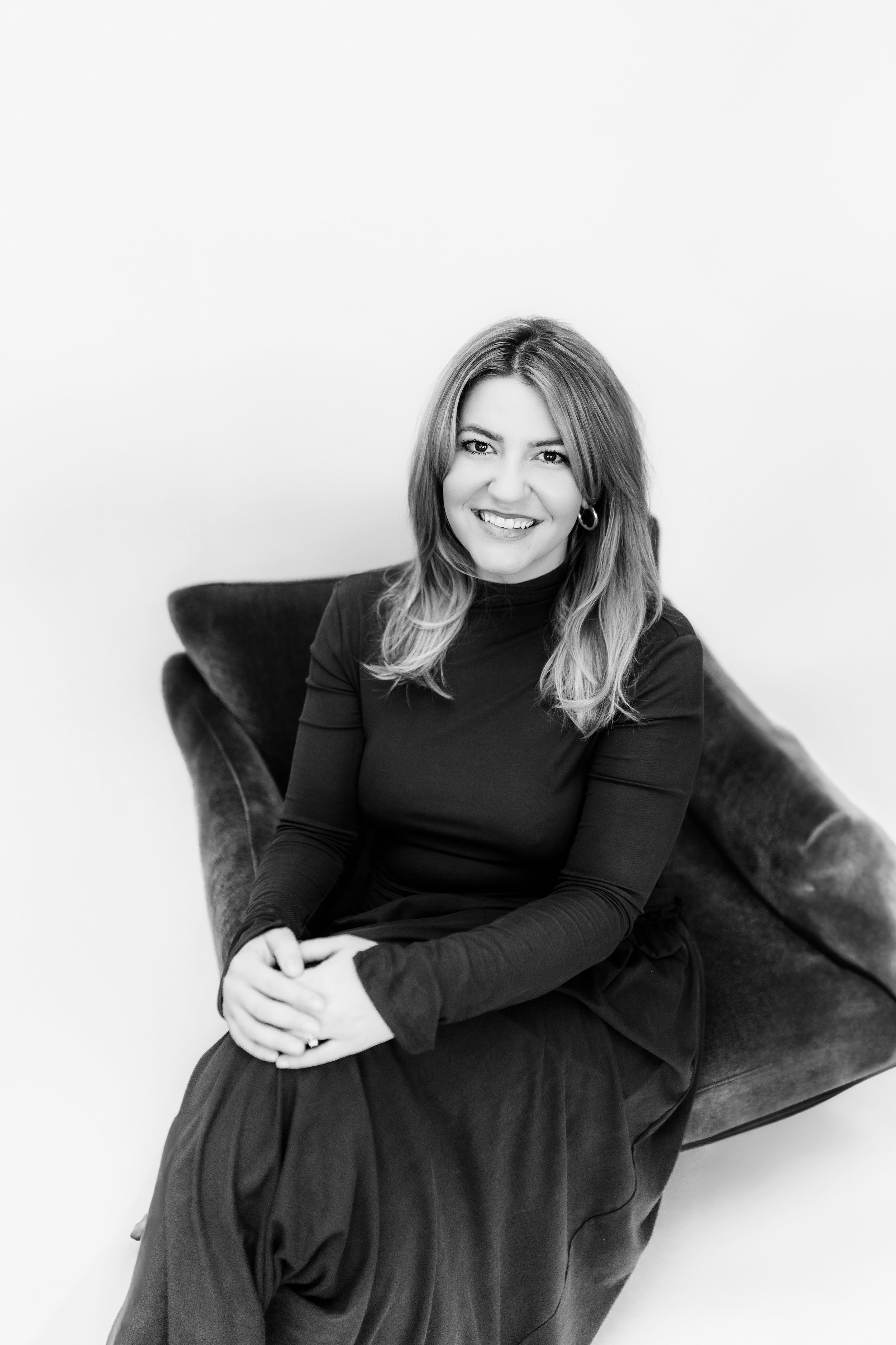 Black and white photo of a woman with shoulder-length hair, smiling, sitting on a velvet armchair, wearing a long-sleeve turtleneck and a long skirt.