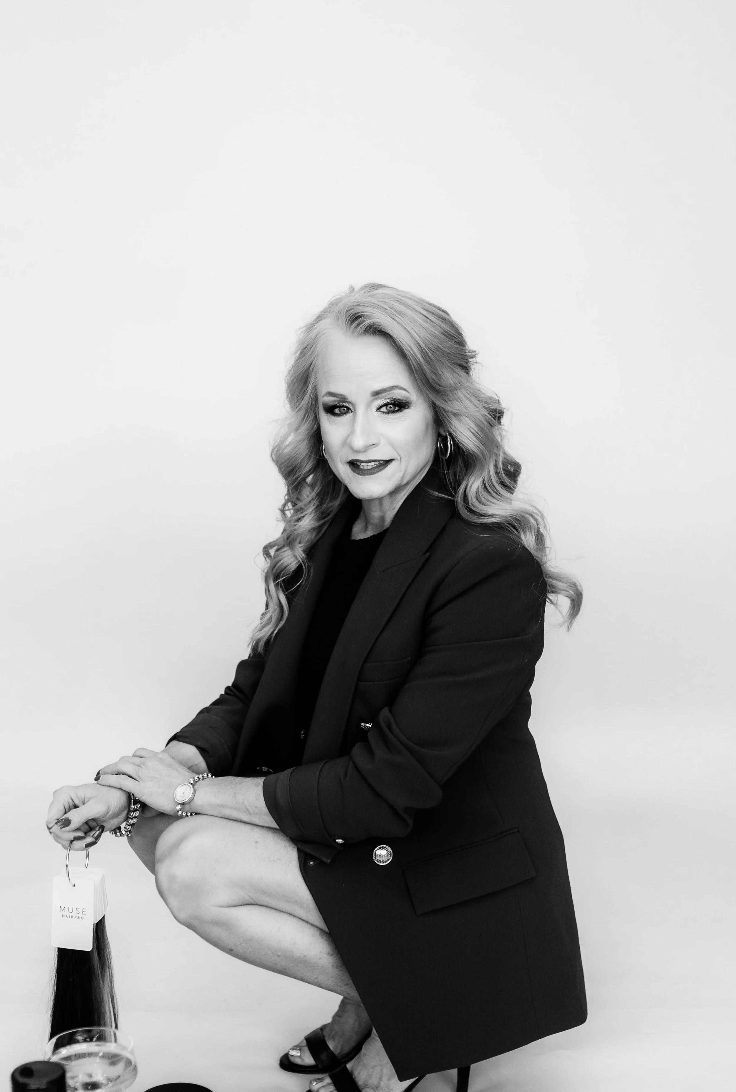 Black and white portrait of a woman with wavy blonde hair, wearing a blazer, sitting with one knee up, smiling slightly, holding a keychain with a hair extension, and wearing jewelry, against a plain background.