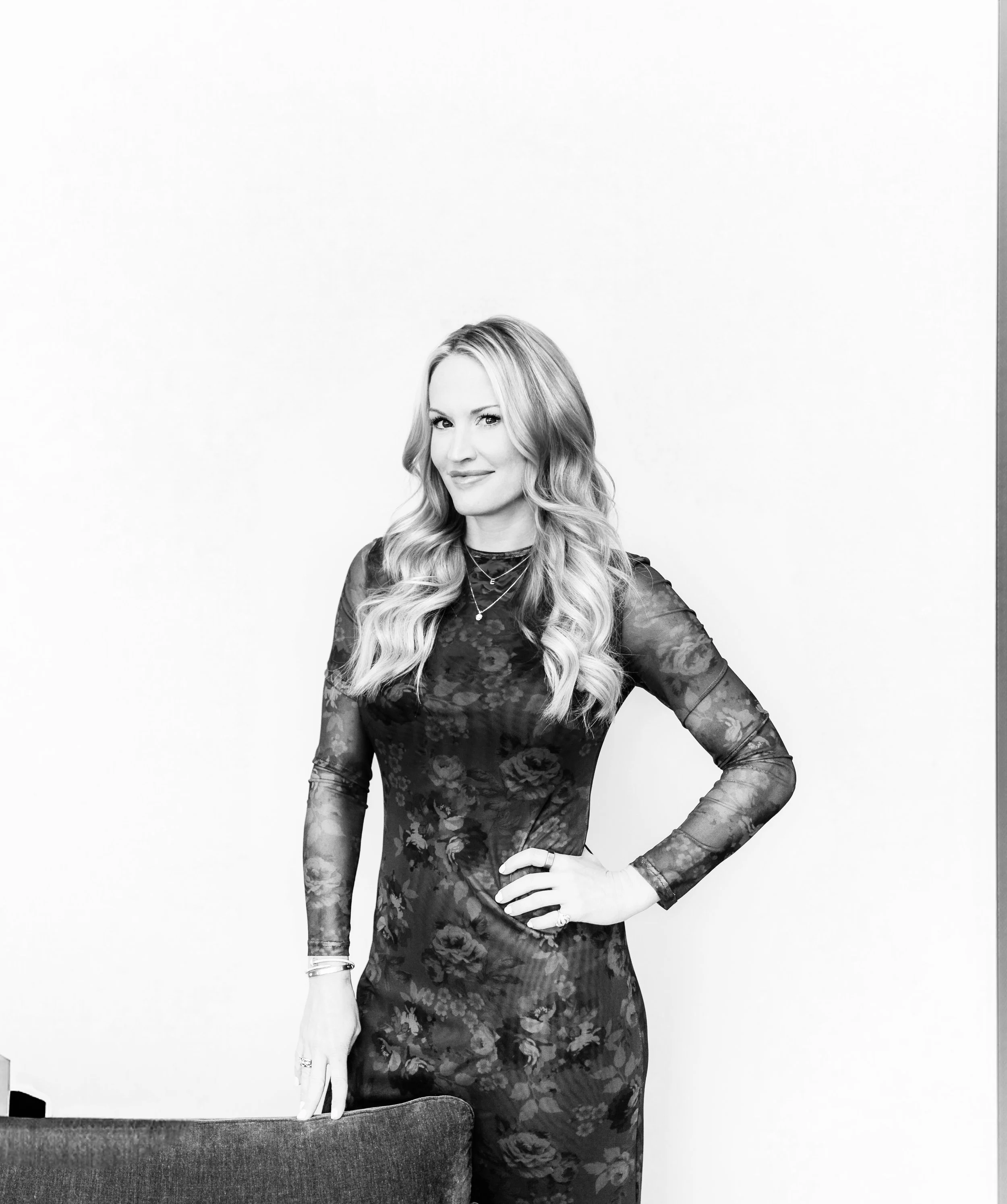 Black and white photo of a woman with long wavy hair wearing a floral dress, standing next to a chair, against a plain background.