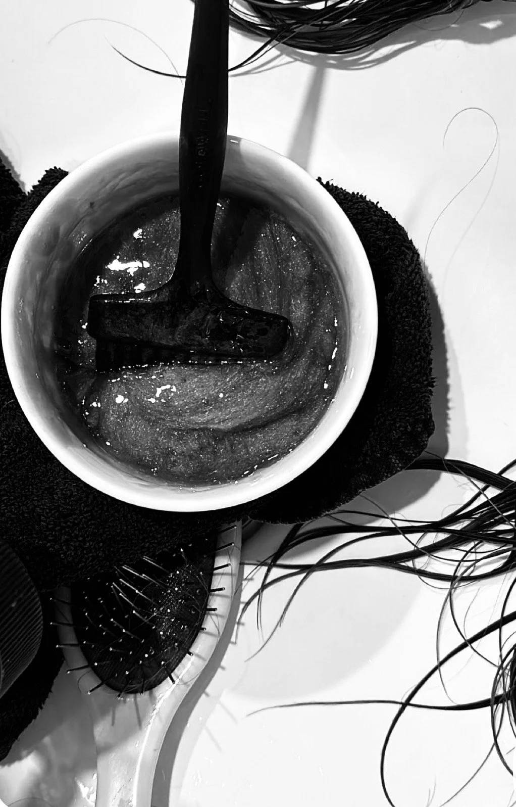 A black and white photo of a hairbrush with black bristles, a cup with hair dye and a black applicator brush inside, surrounded by hair strands, and a towel underneath the cup.