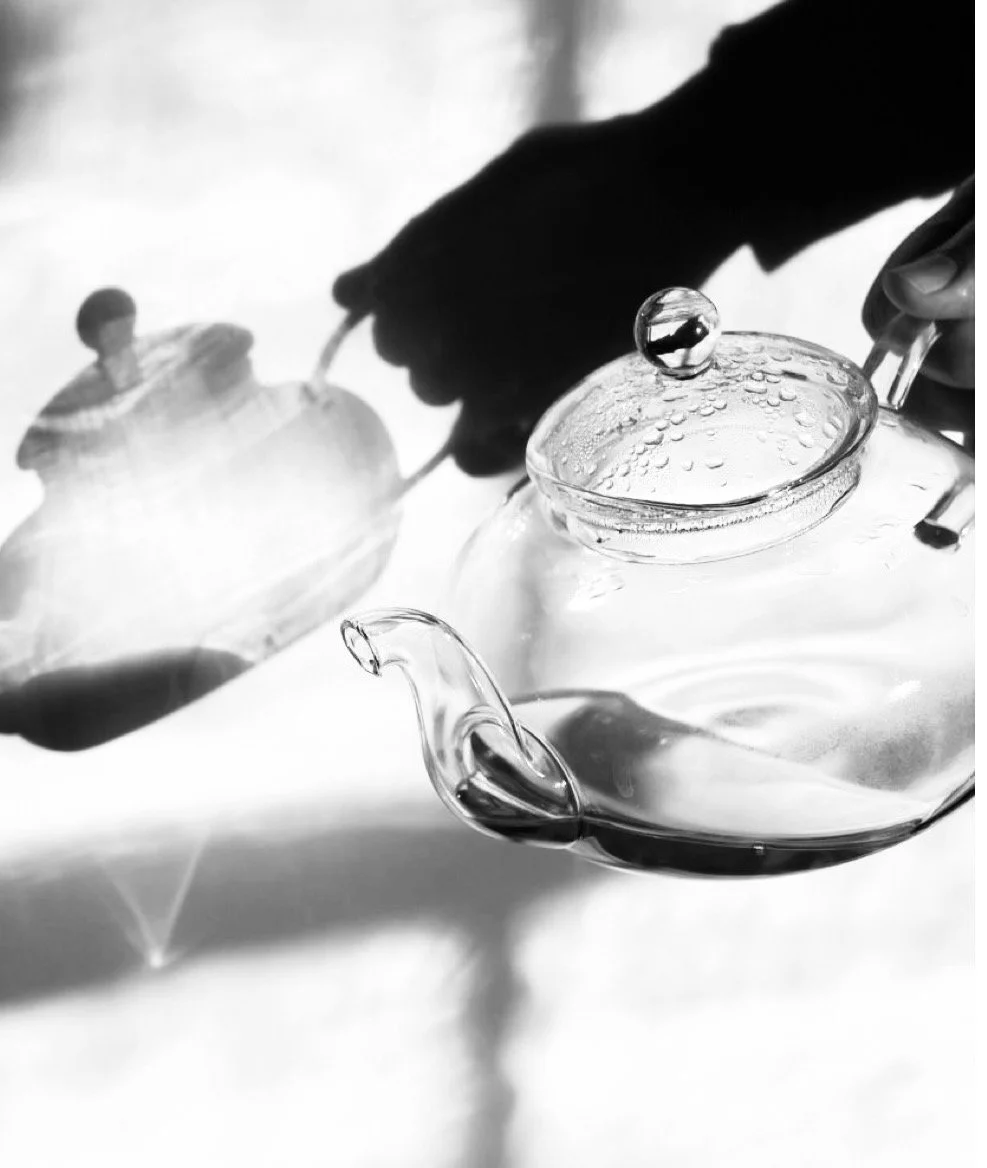 Black and white close-up of a person's hand pouring liquid from a glass teapot into a cup.